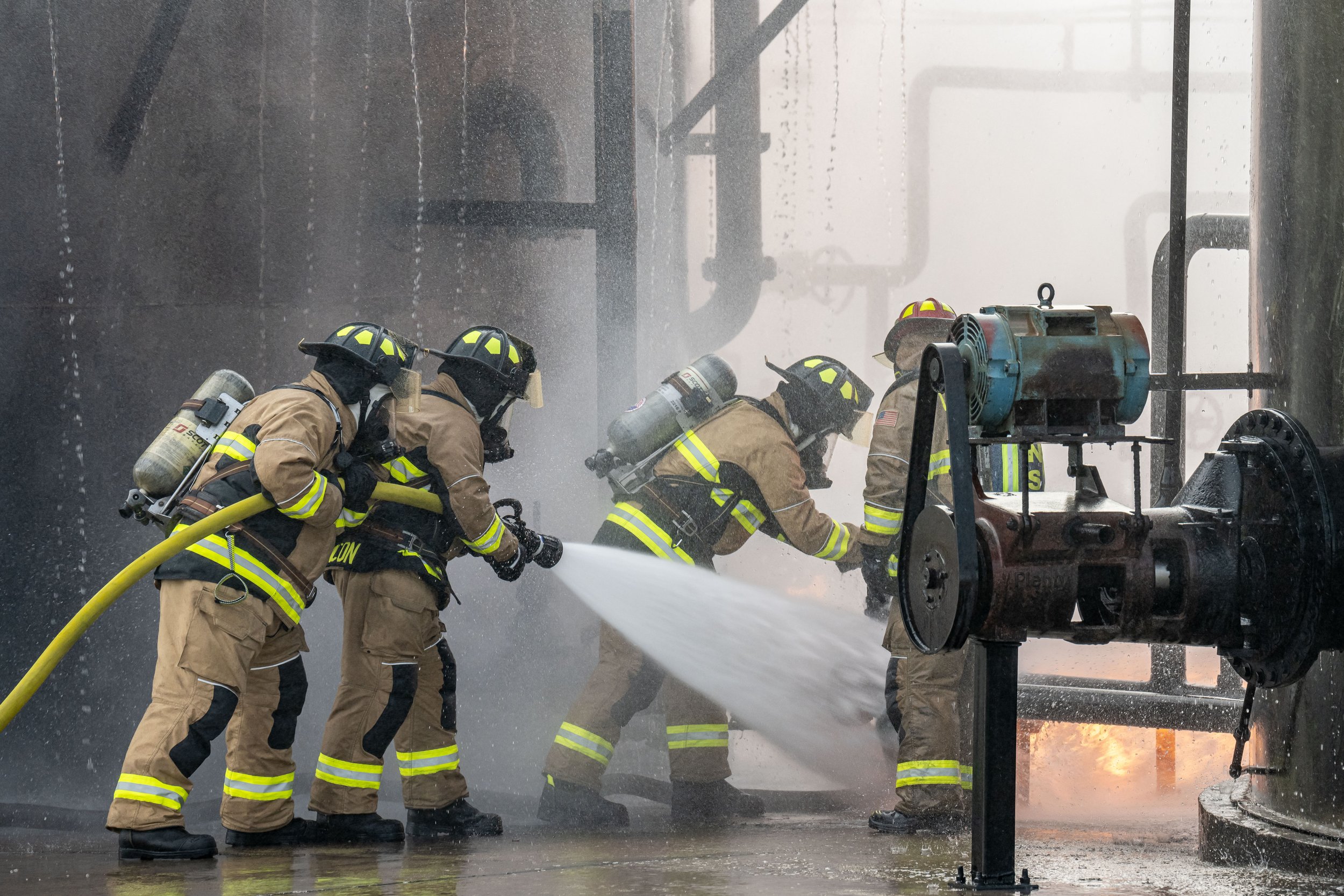 Firefighters in full gear using a hose to spray water on a fire inside an industrial facility.