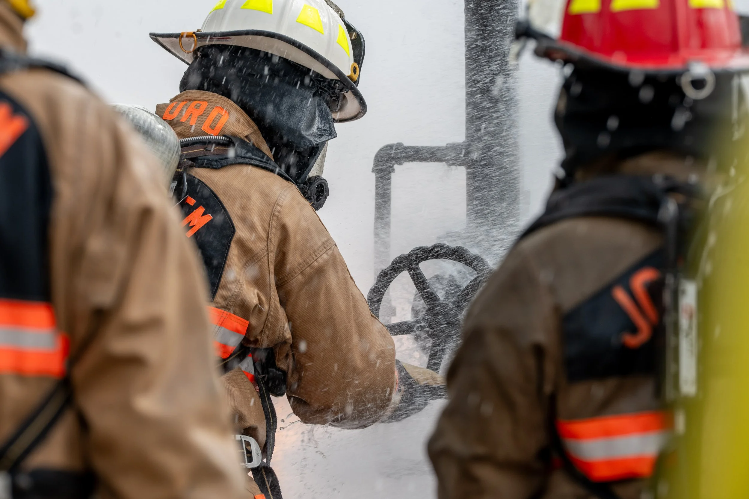 Three firefighters in protective gear and helmets using a hose to spray water or foam.