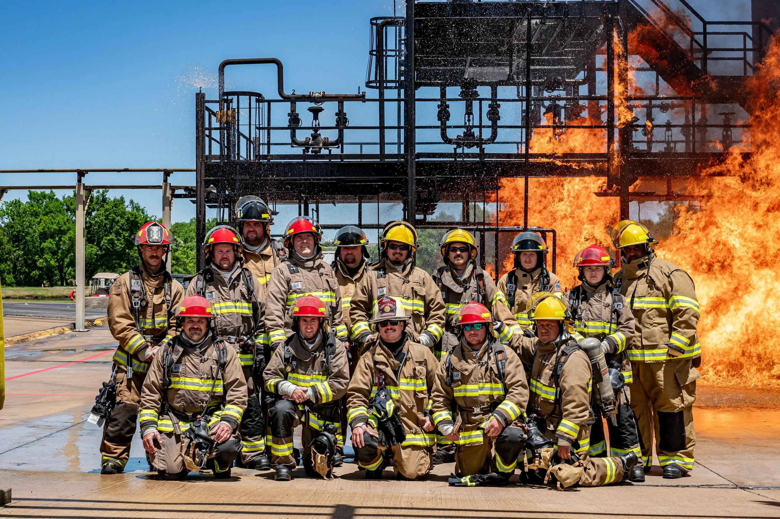 Group of firefighters in uniform posing in front of a large fire at an industrial site.