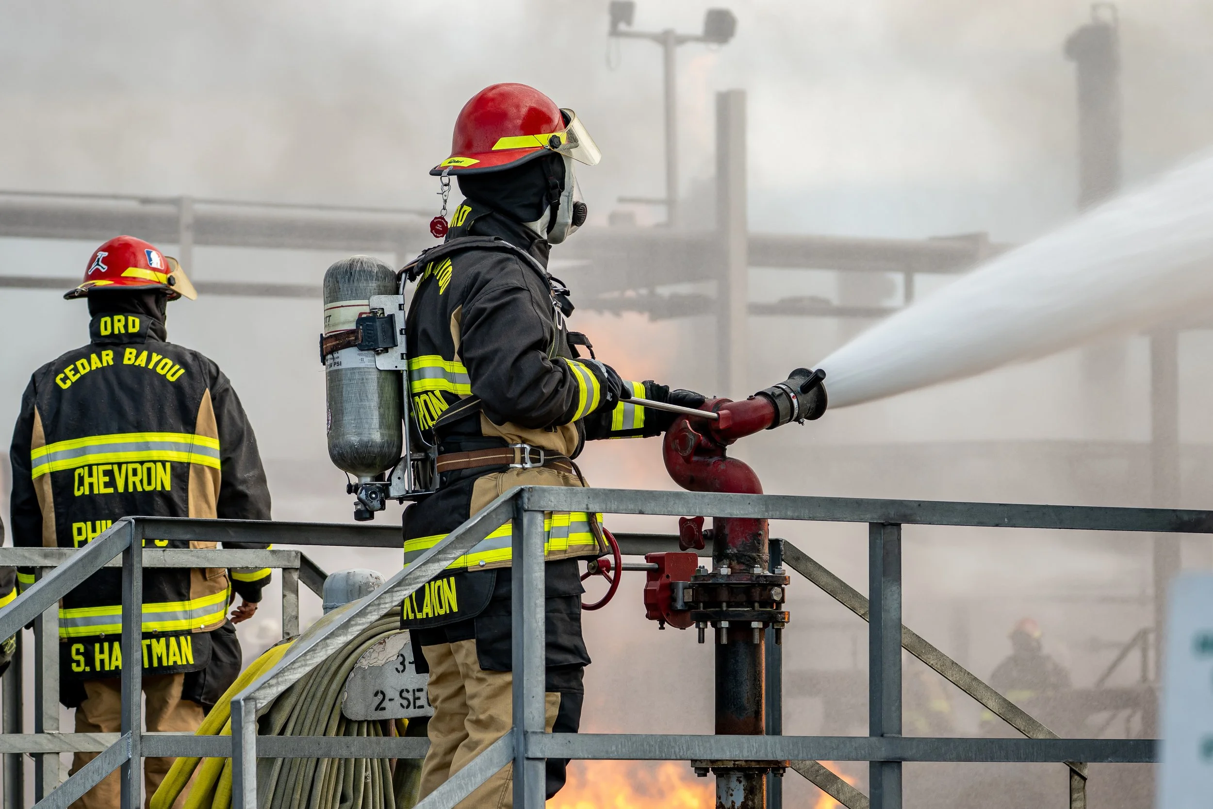 Firefighters in protective gear operating a fire hose to extinguish a fire on an industrial structure.