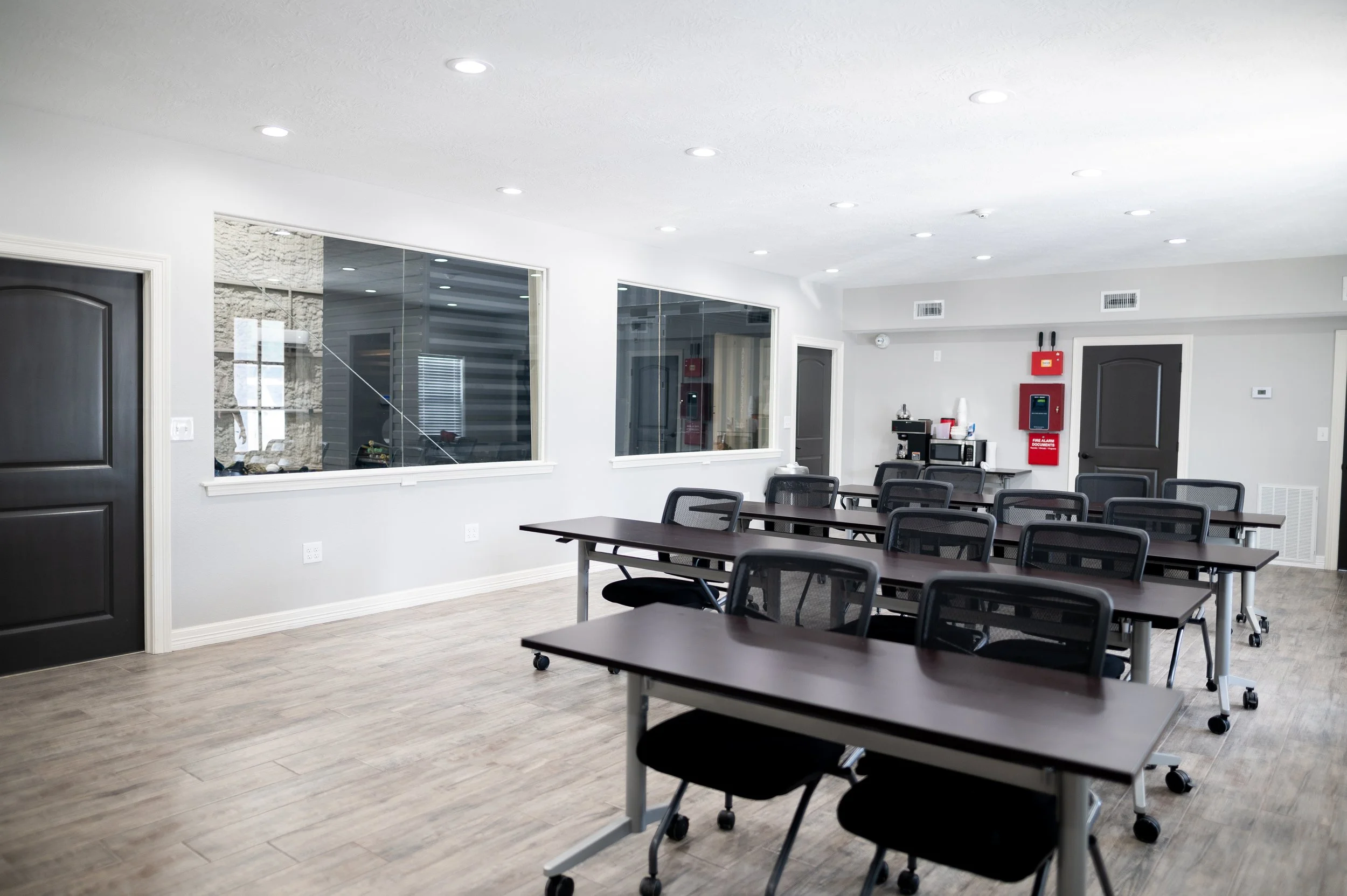 Empty classroom or meeting room with multiple tables and chairs, windows, and fire safety equipment on the wall.