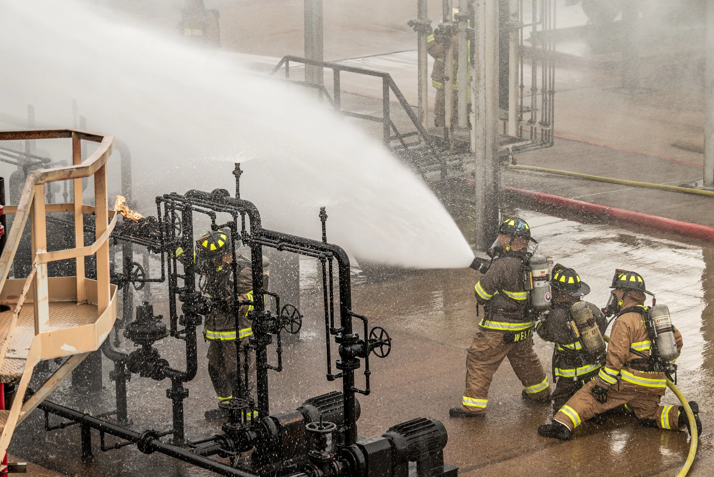 Firefighters in protective gear spraying water on a fire at an industrial site with pipes and machinery.