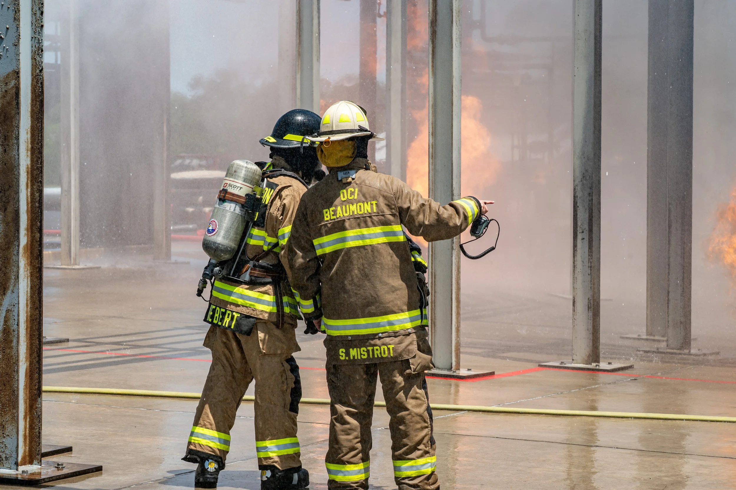 Three firefighters in full gear working near a fire with flames visible in the background.
