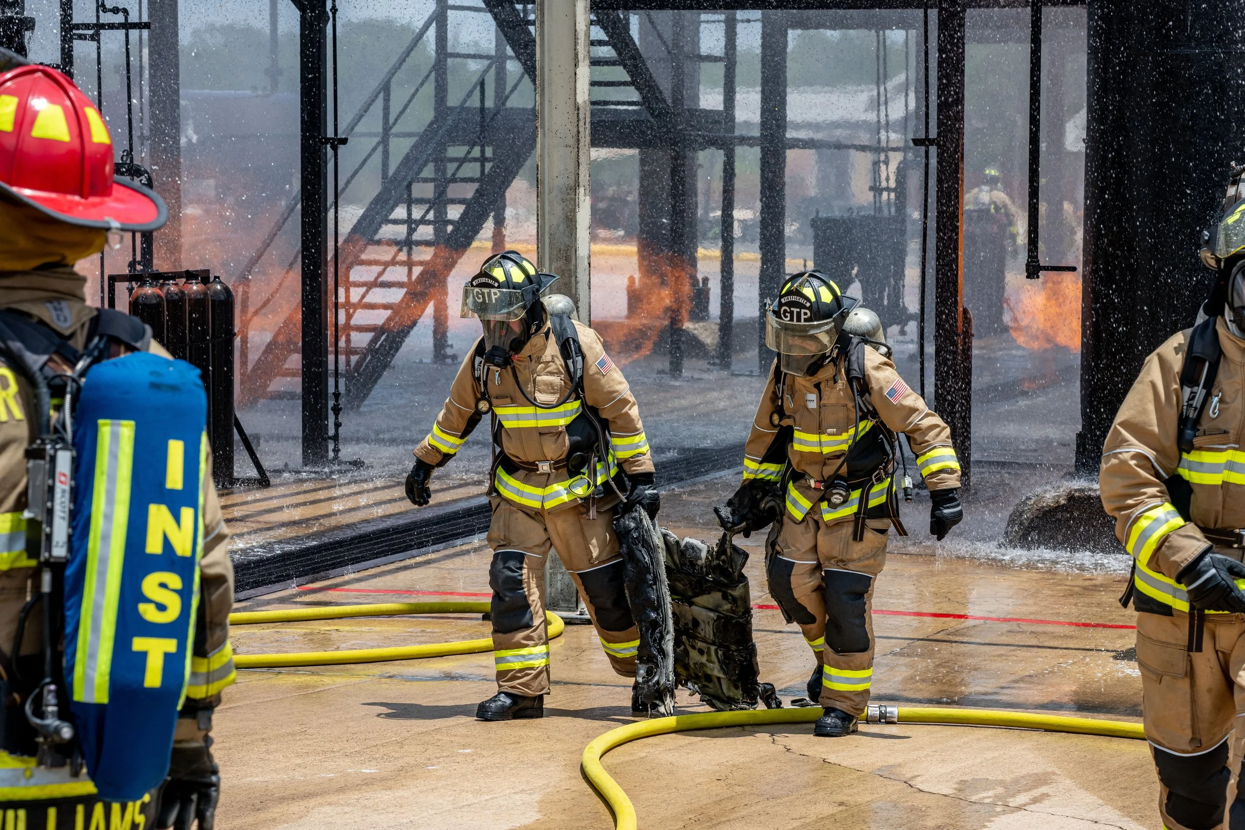 Firefighters in full gear, including helmets and protective suits, respond to a fire at a building with flames and smoke visible in the background, using hoses to extinguish flames.