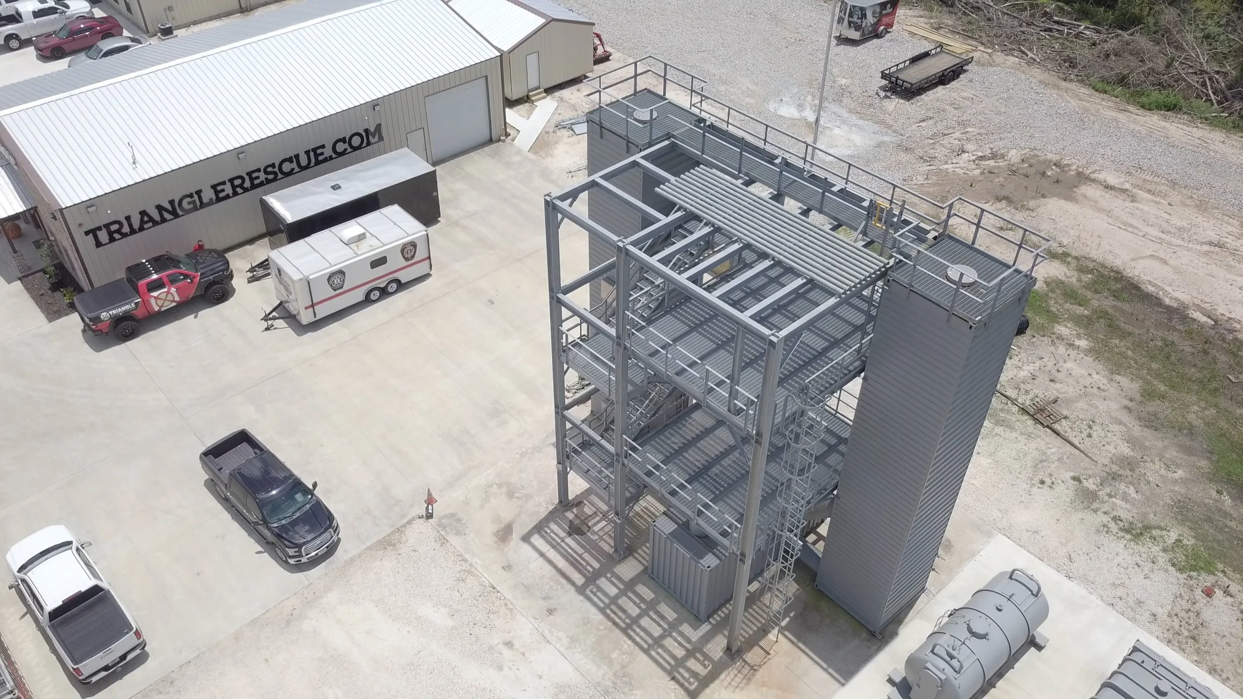An aerial view of a rescue operation site showing a large metal rescue tower, vehicles, trailers, and a building with the text 'TRIANGLE RESCUE.COM' on it.