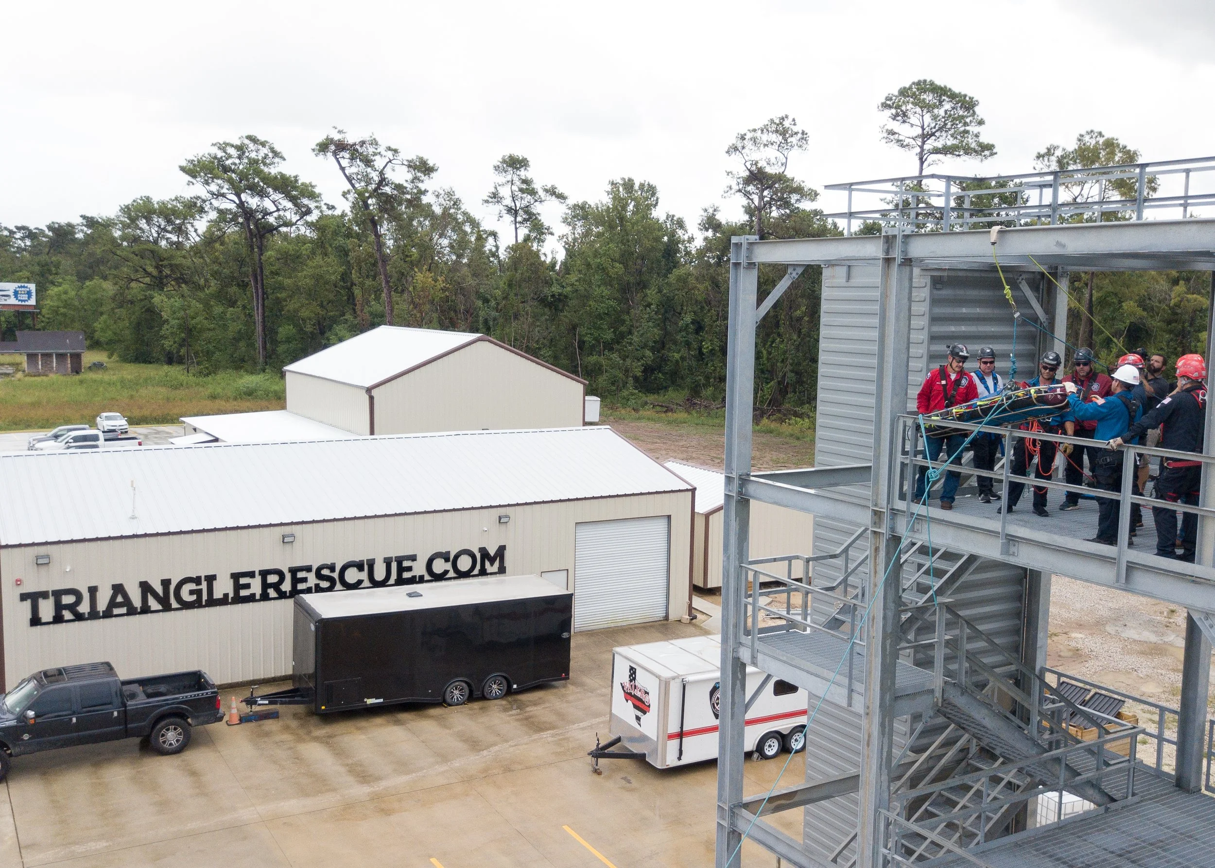 Group of rescue workers wearing safety helmets and harnesses standing on an outdoor metal staircase, preparing equipment, with trees in the background and a building with signage reading 'TRIANGLE RESCUE.COM'.