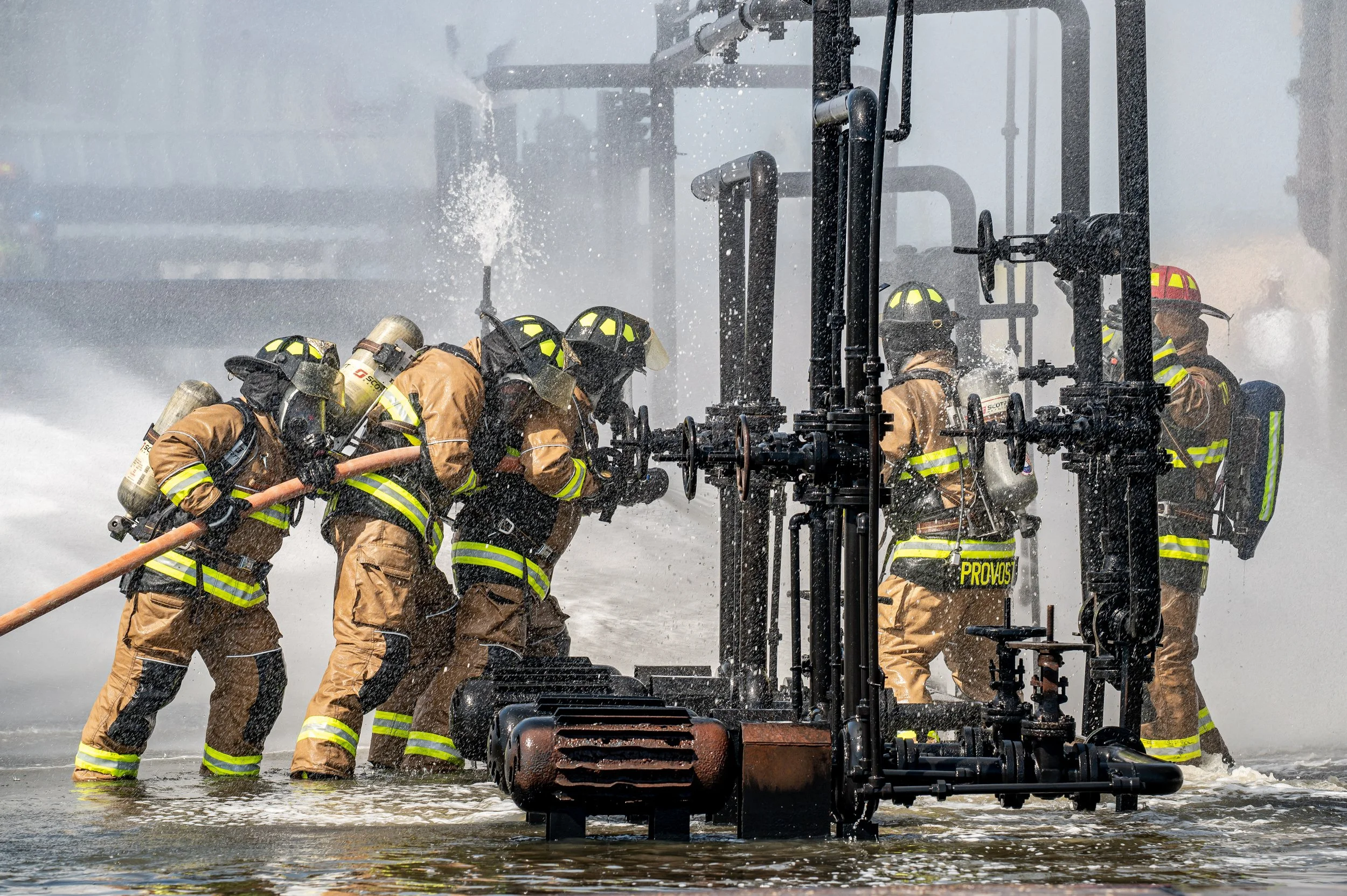 Firefighters in firefighting gear working together to operate a water pump, spraying water and working in water up to their ankles.