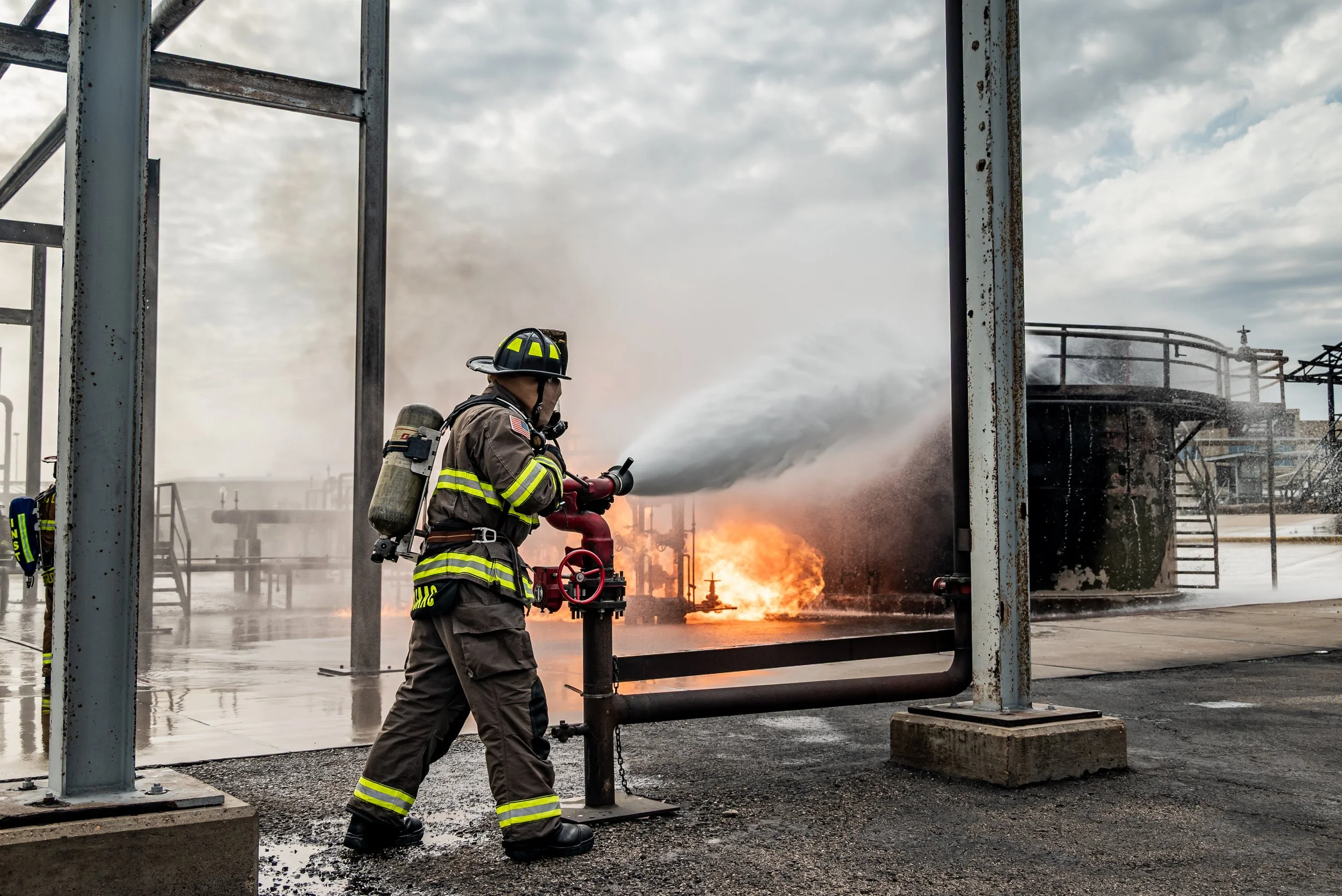 Firefighter in turnout gear and helmet operating a fire hose, spraying water at a fire on an industrial site with metal structures and a cloudy sky.