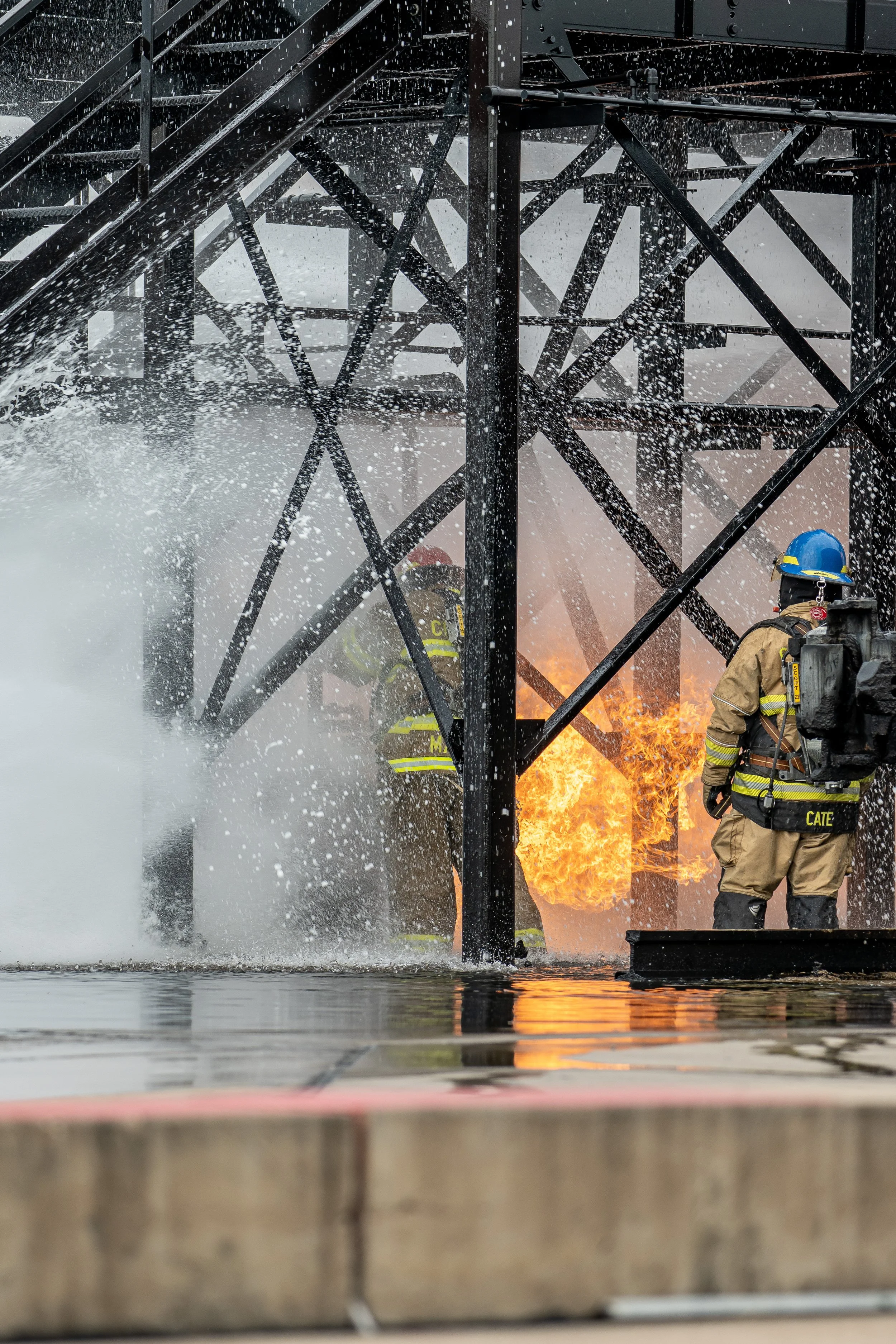 Firefighters in protective gear fighting fire and smoke on a metal structure, with flames visible in the background.