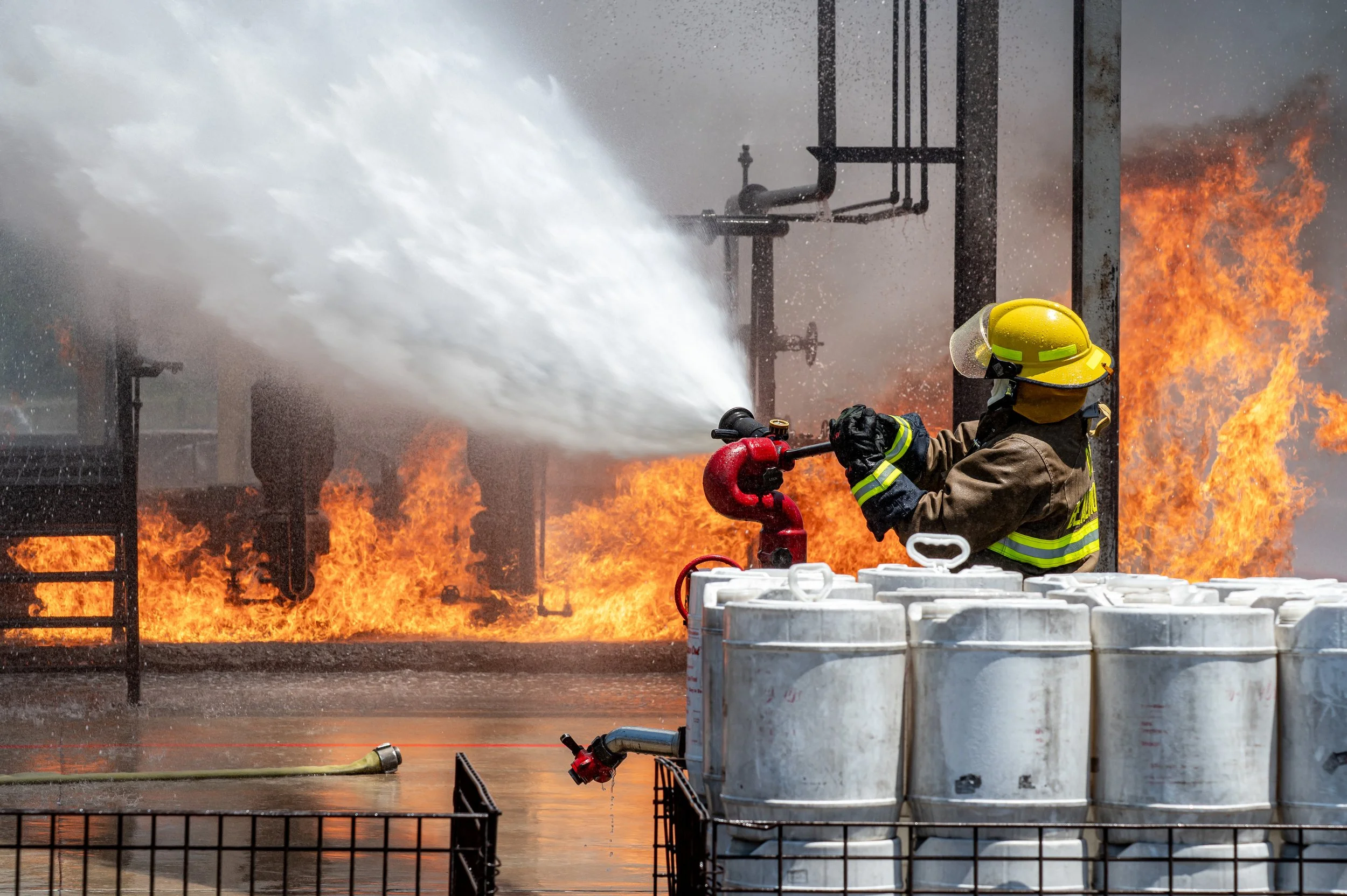 Firefighter in protective gear battling a fire with a hose, with flames and smoke in the background.