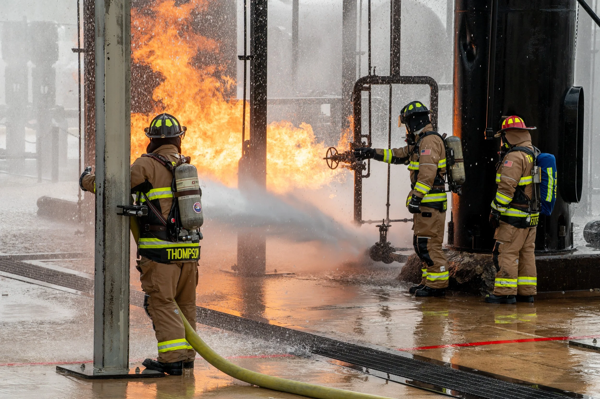 Four firefighters in full gear working together to control a fire involving industrial equipment, with flames and water spray visible.