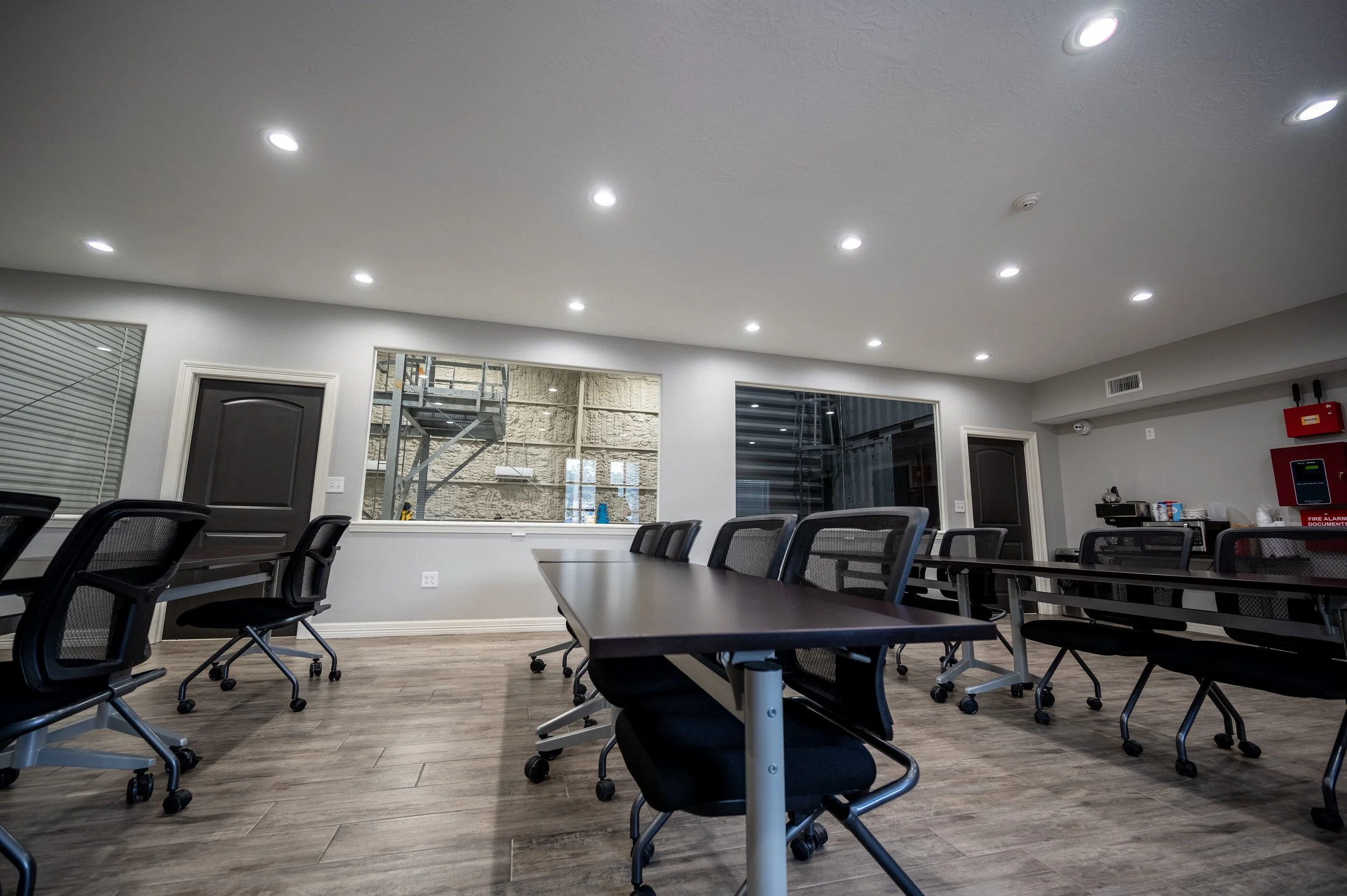 Empty conference room with wooden floor, black chairs, and rectangular tables; white walls with large windows and ceiling lights.