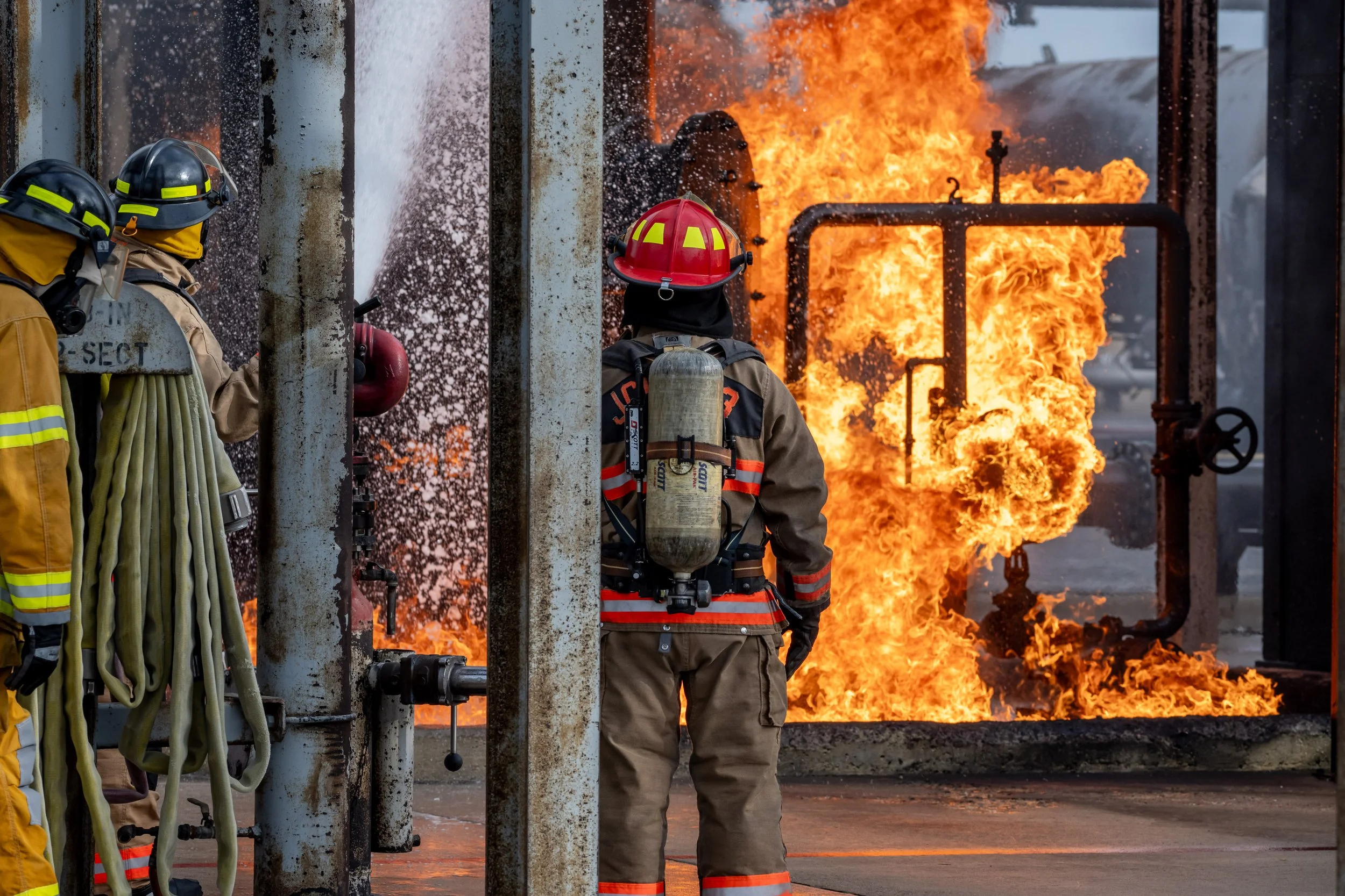 Three firefighters in protective gear working near a large blaze and flames in an industrial setting.