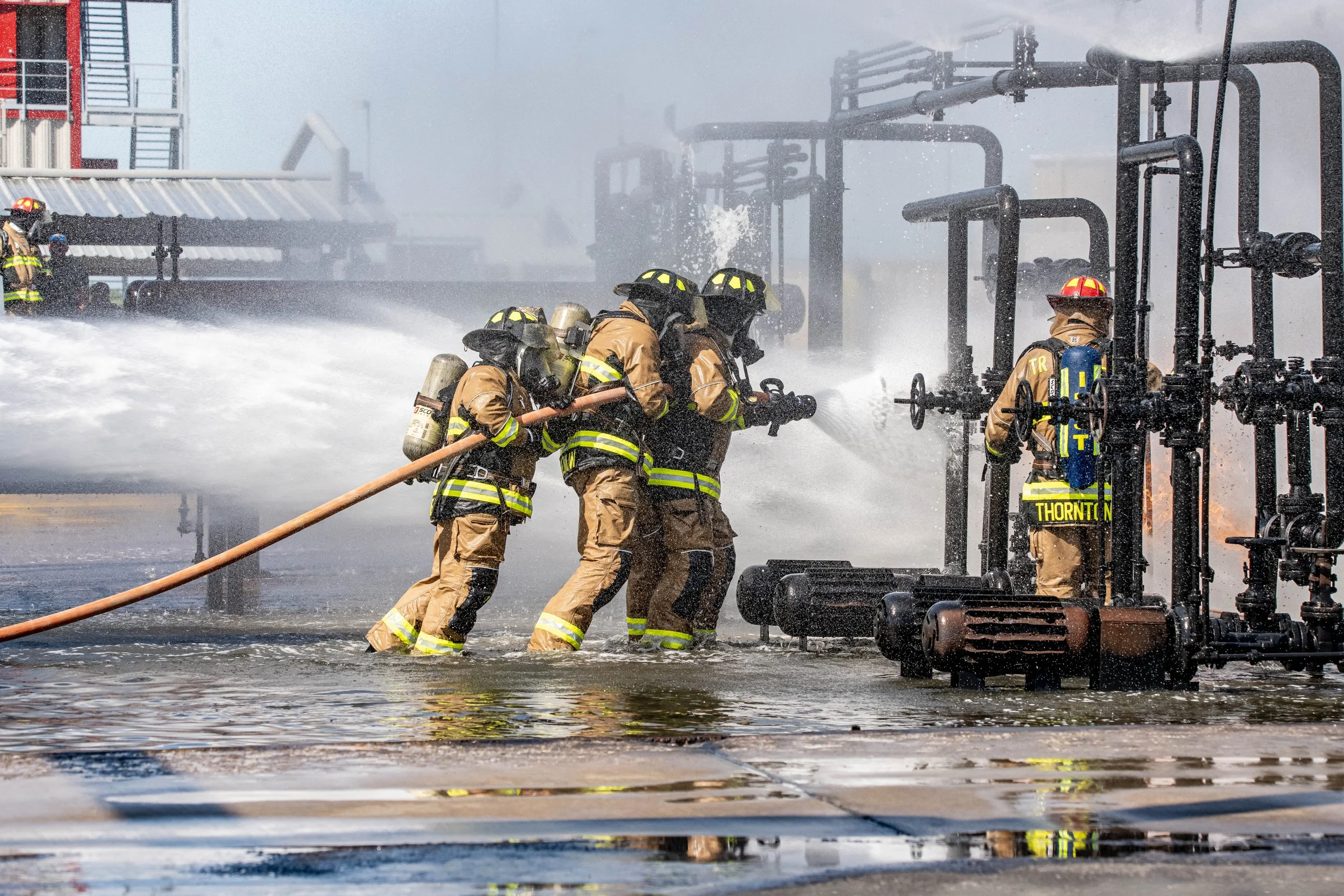 Firefighters in full gear training or responding to a fire at an industrial site, with water spraying from a hose and chemicals or water vapor in the background.