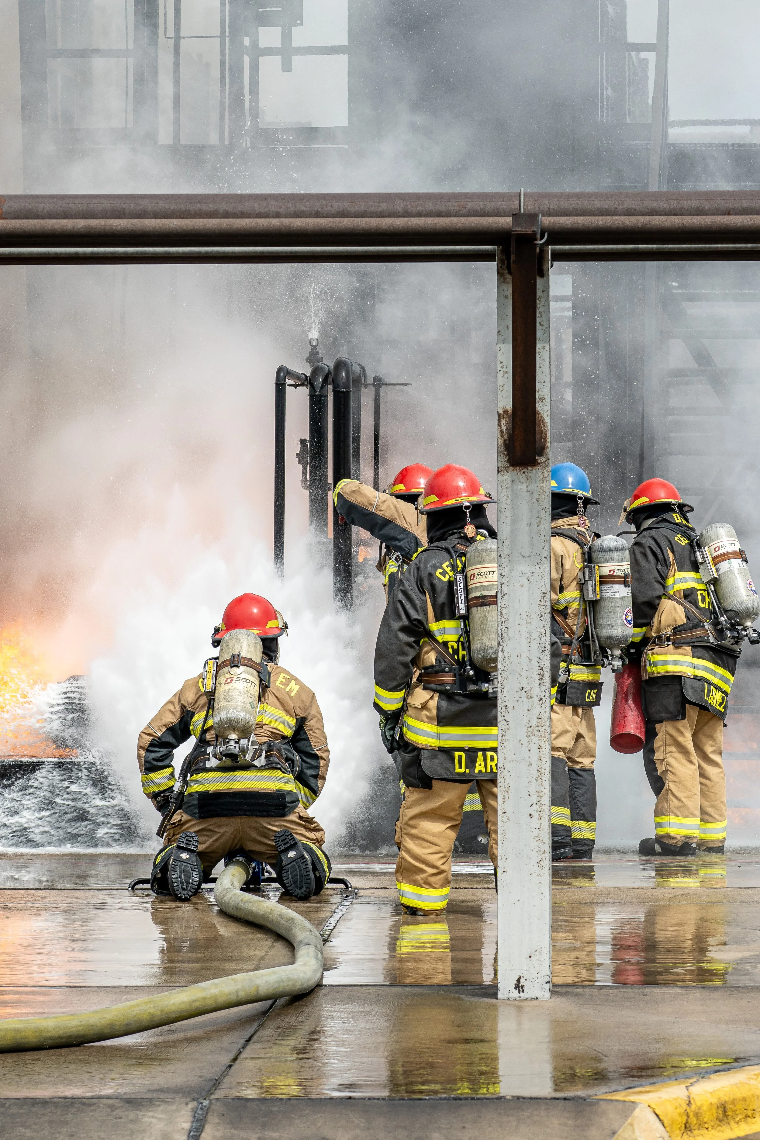 Firefighters battling a fire with hoses, surrounded by smoke and flames in an industrial setting.