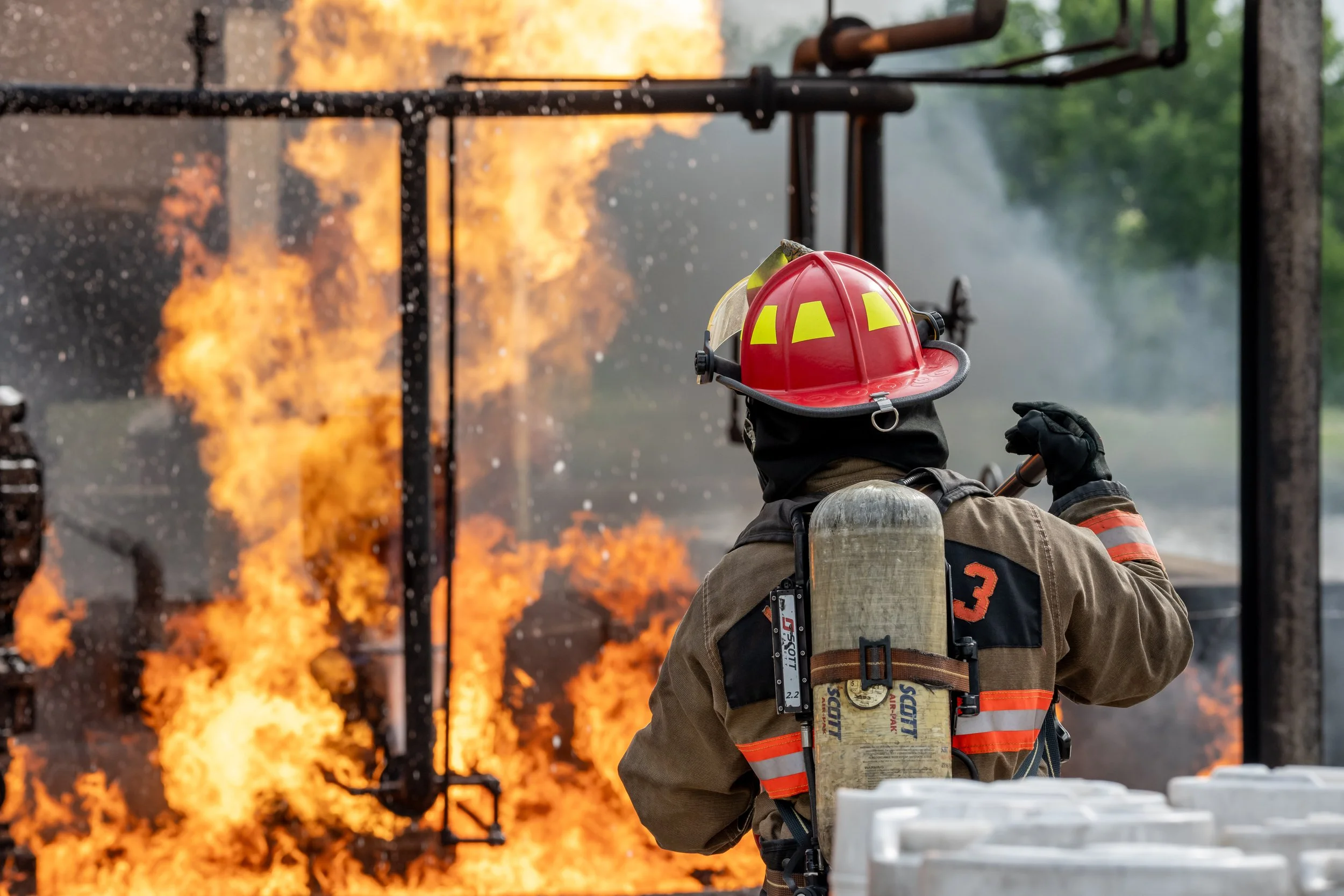 Firefighter in protective gear and helmet using a tool to combat a large fire on a structure.