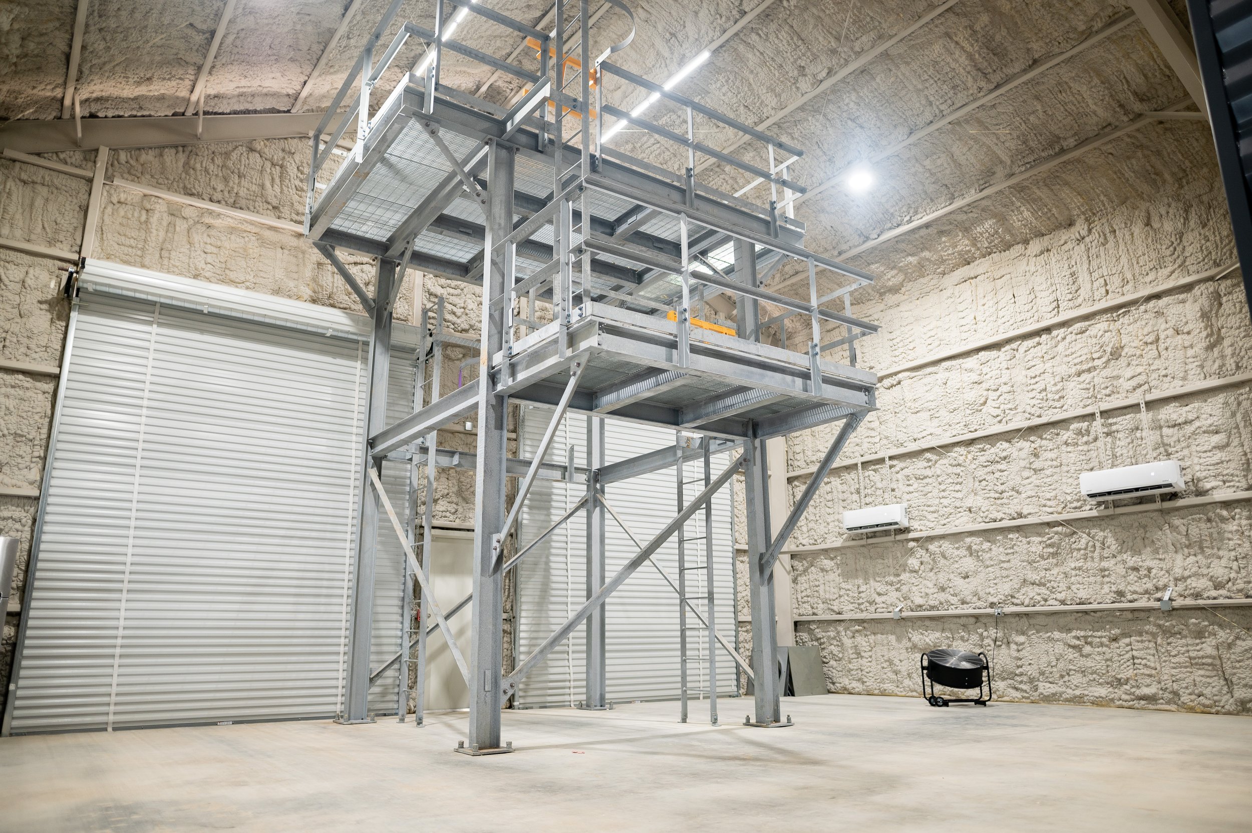 Empty industrial or maintenance warehouse space with a metal scaffolding structure, insulated walls, a roll-up door, and a small black fan or heater on the floor.