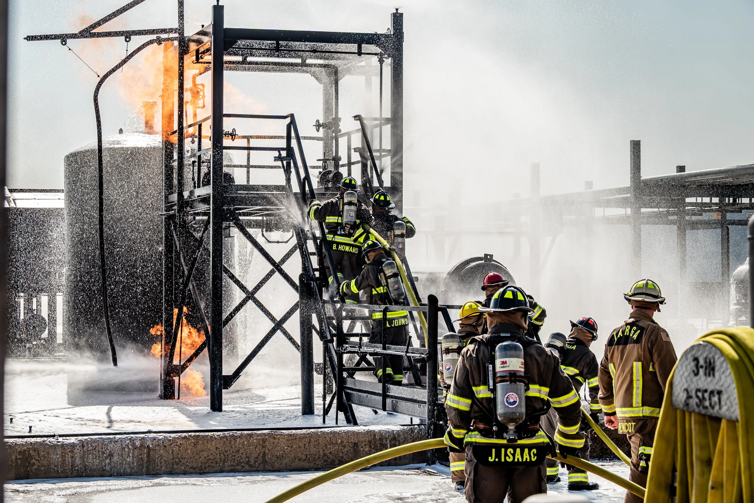 Firefighters combat a fire at an industrial site with flames visible and water spraying from hoses.