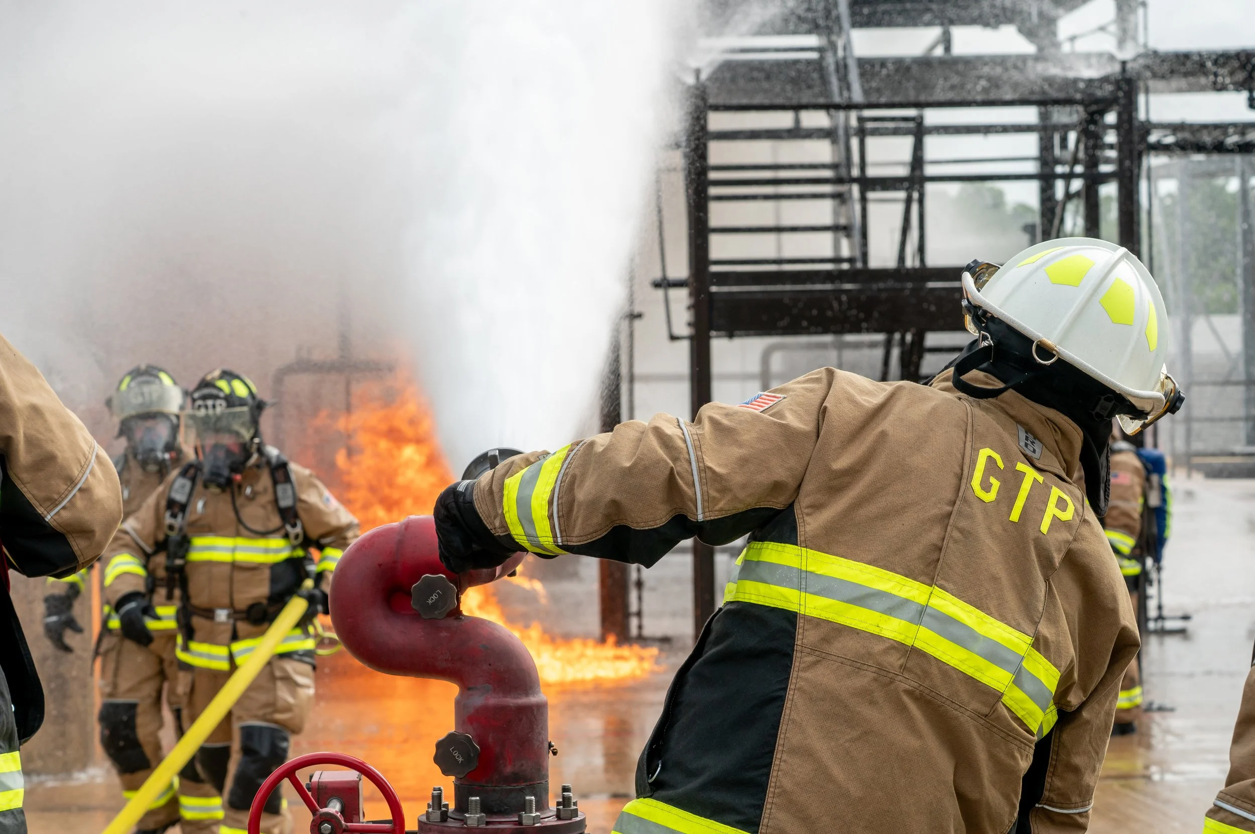 Firefighters in protective gear are fighting a fire at an industrial site, with one firefighter adjusting a red fire hose, and flames and smoke visible in the background.