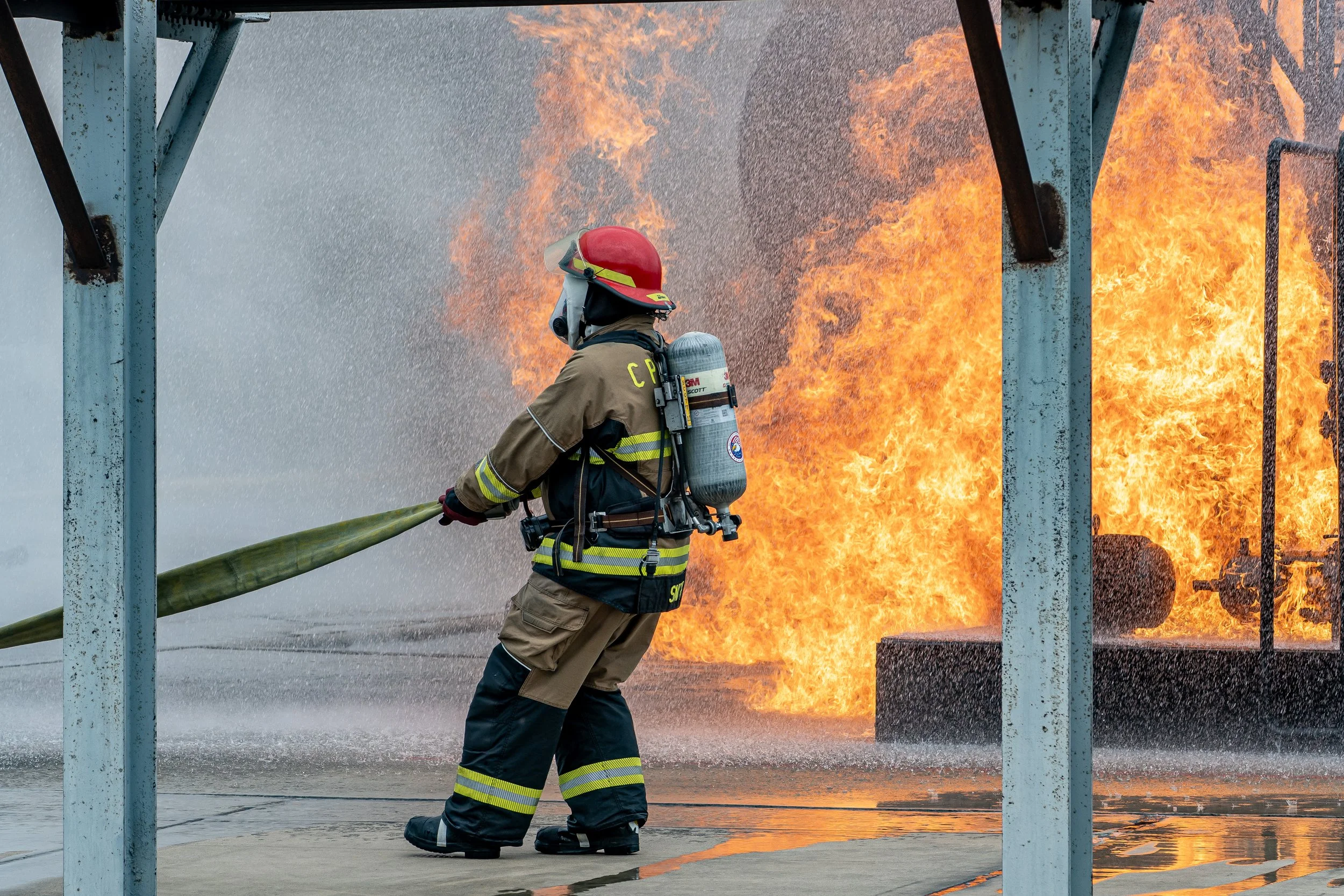 Firefighter in protective gear spraying water on a building fire with large flames in the background.