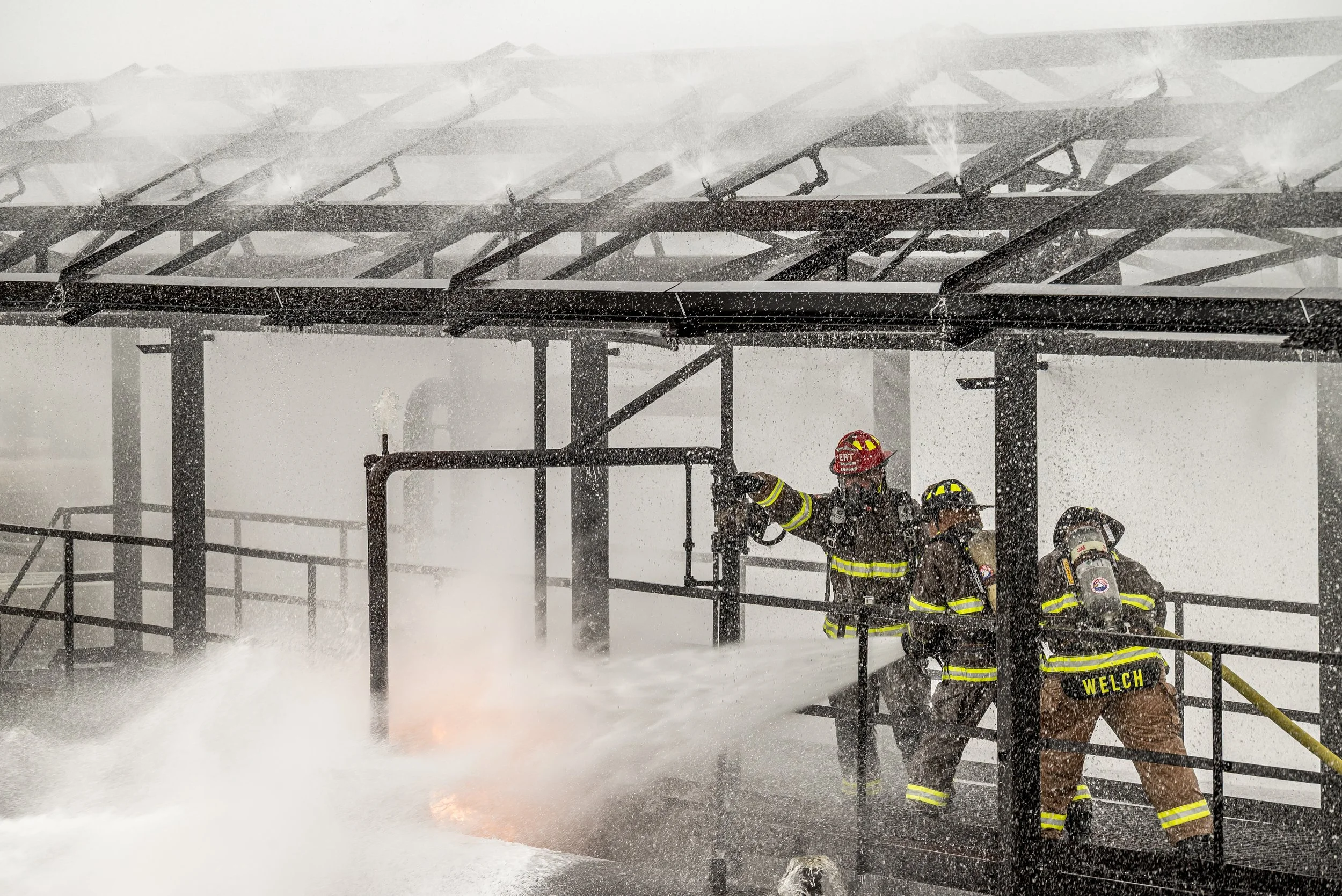 Firefighters fighting a blaze on a smoke-filled, rainy rooftop with spraying water.