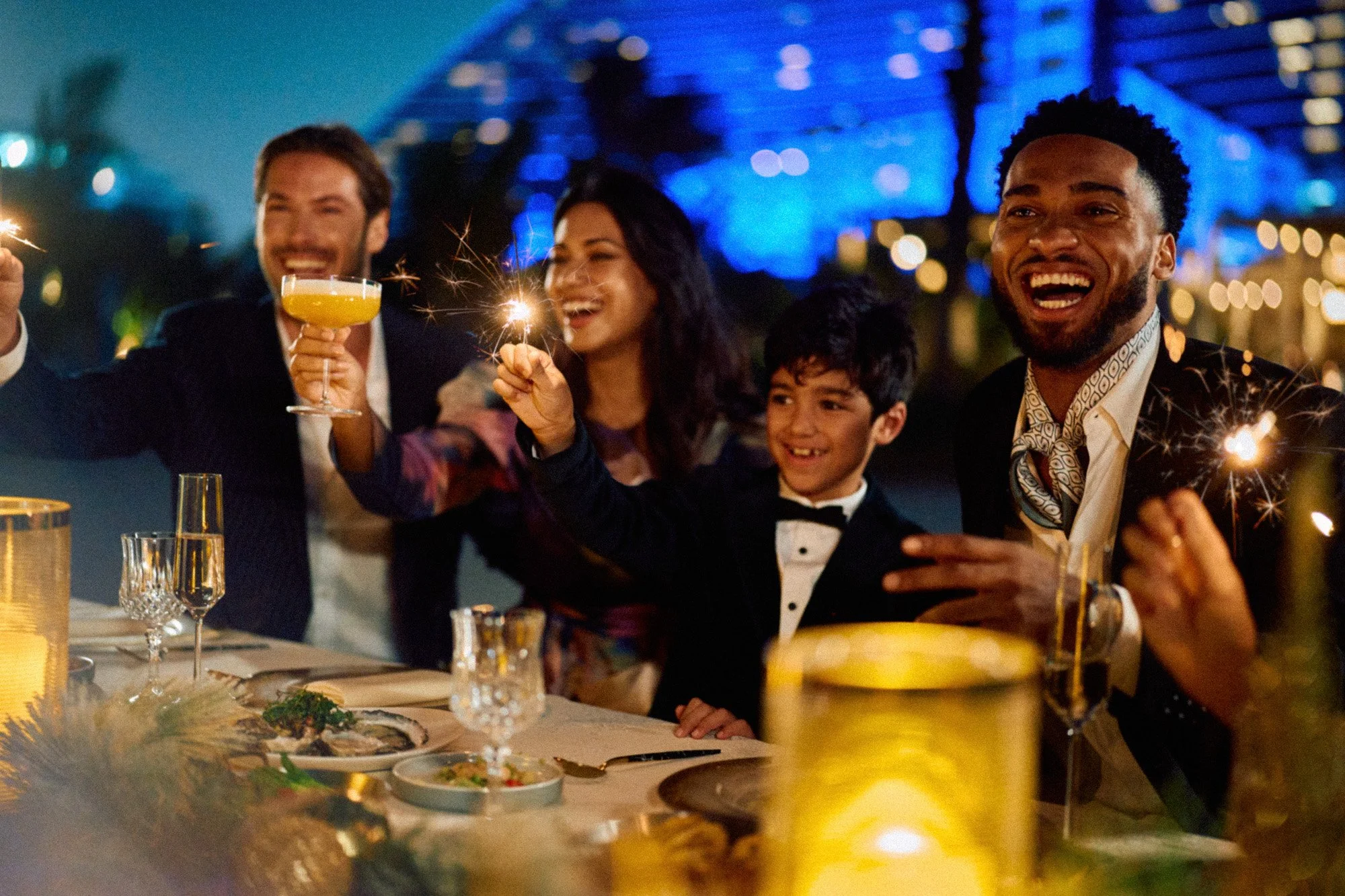 A group of people celebrating at a dinner party, holding sparklers and drinks, with a festive outdoor setting and colorful lights in the background.