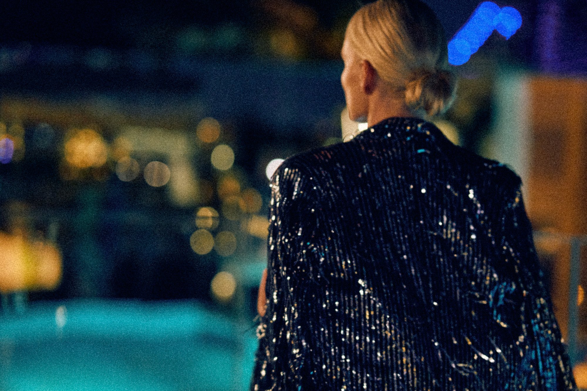 Woman with blonde hair in a bun, wearing a sparkling black sequin jacket, standing outdoors at night with city lights in the background.