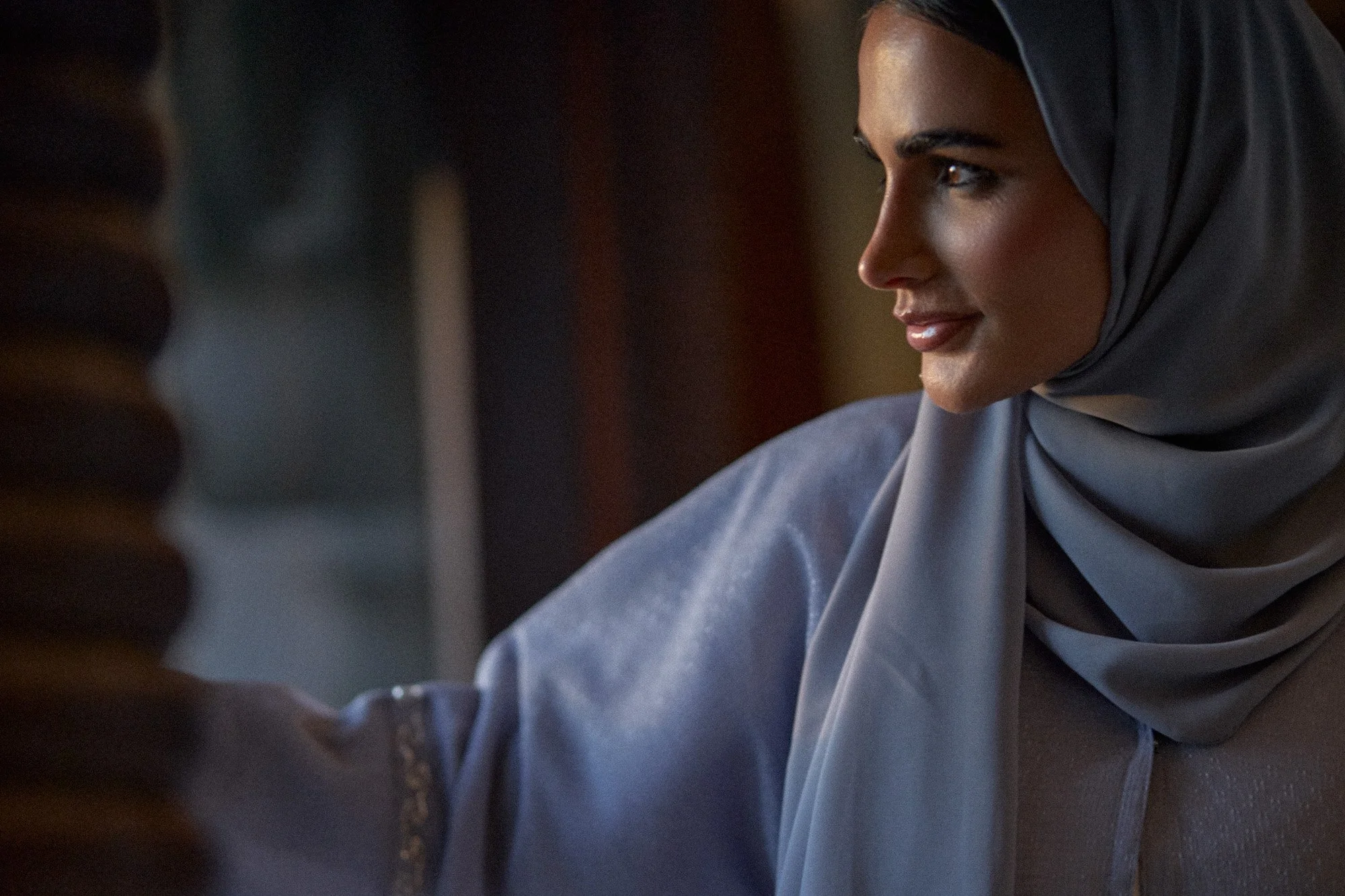 A woman wearing a gray hijab and gray top looking to the side with a soft smile in a warmly lit room.