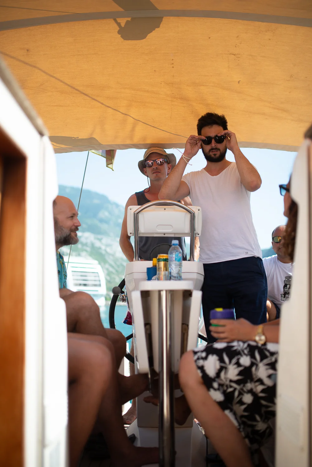A group of people on a boat enjoying sunny weather, with some wearing sunglasses and hats, in a scenic outdoor setting with mountains in the background.