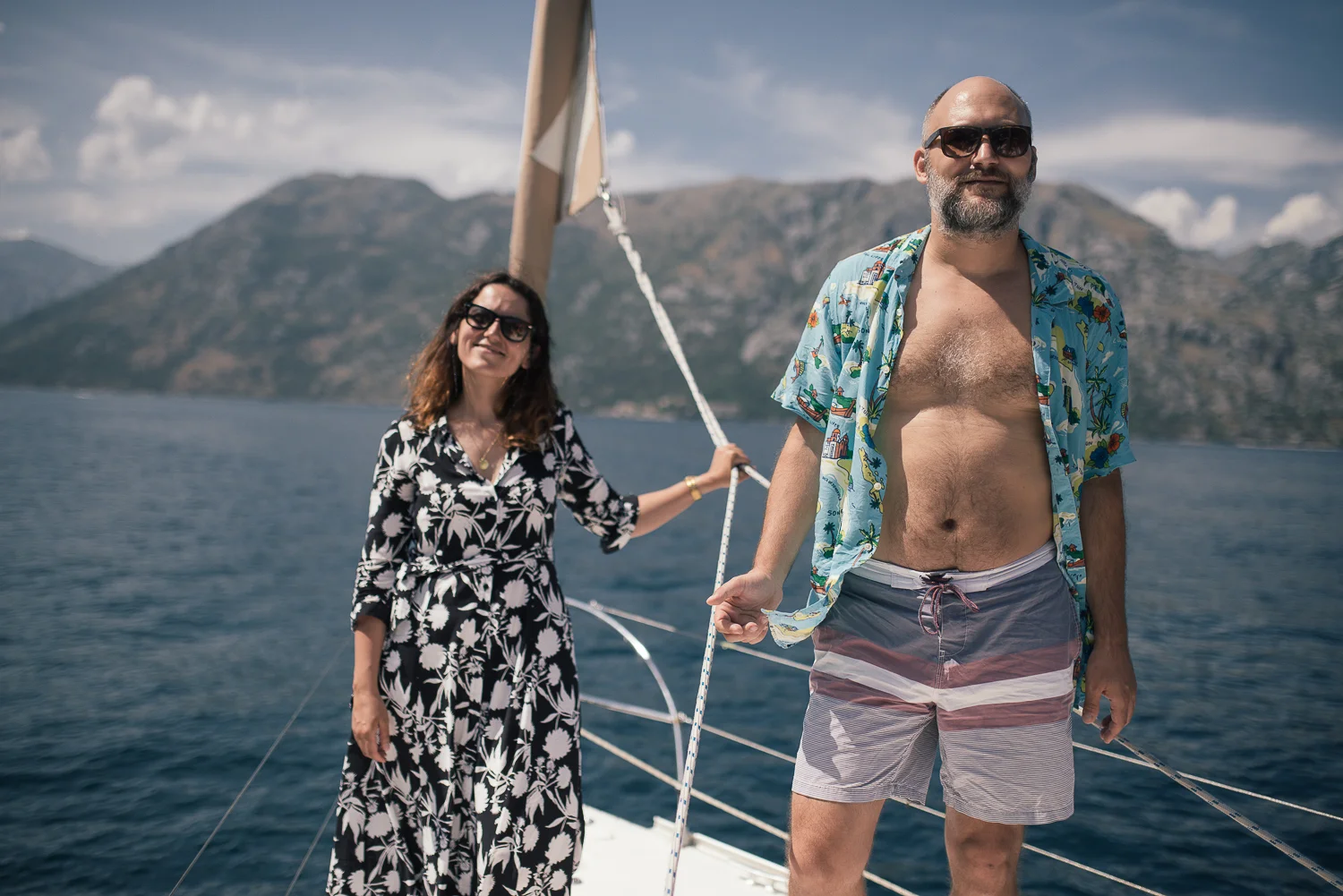 A man and woman are on a boat with a mountainous landscape in the background. The man is wearing sunglasses, an open blue Hawaiian shirt, and striped swim trunks. The woman is wearing sunglasses and a black and white floral dress.
