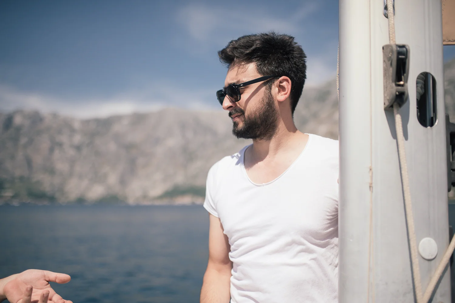 Man with dark hair, beard, and sunglasses standing on a boat near the water, with mountains and cloudy sky in the background.