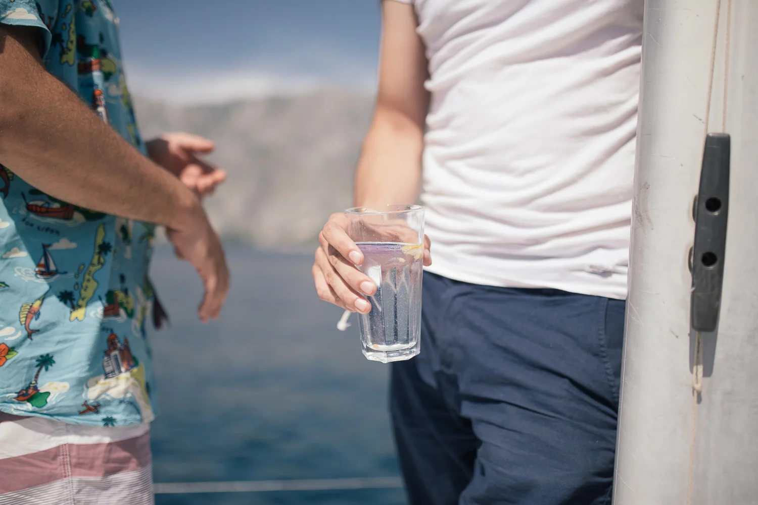 Person holding a glass of water on a boat or near water, with another person partially visible in swimwear.