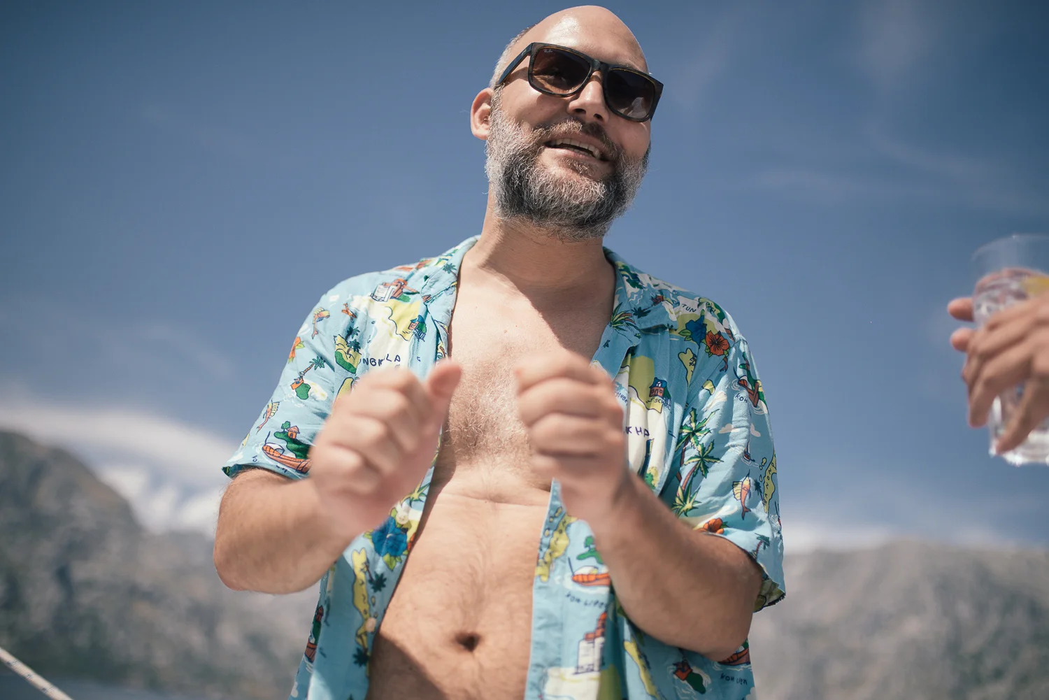 A man with a beard and sunglasses enjoying the outdoors, wearing a colorful Hawaiian shirt open at the front, near a body of water with mountains in the background.