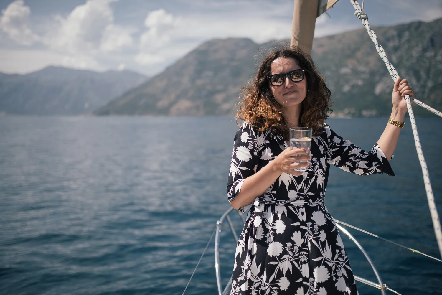 Woman with curly hair wearing sunglasses and a black and white floral dress holding a glass of water on a boat with a lake and mountains in the background.