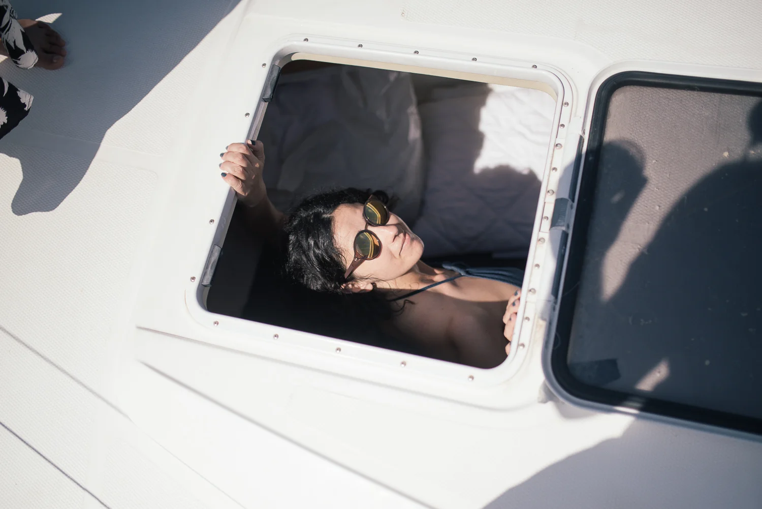 A woman with dark hair and sunglasses is lying down inside a boat hatch, sunbathing on a sunny day.