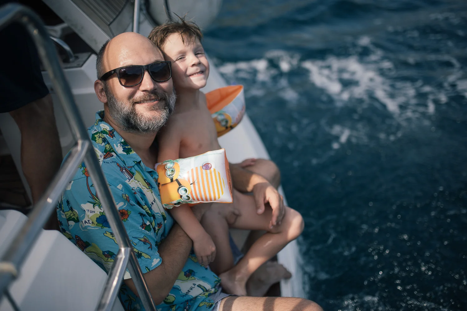A man with sunglasses and a beard smiling, sitting next to a young boy with wet hair on a boat. Both are near the edge, with the ocean visible in the background. The man is wearing a colorful Hawaiian shirt, and the boy has a floatation device.