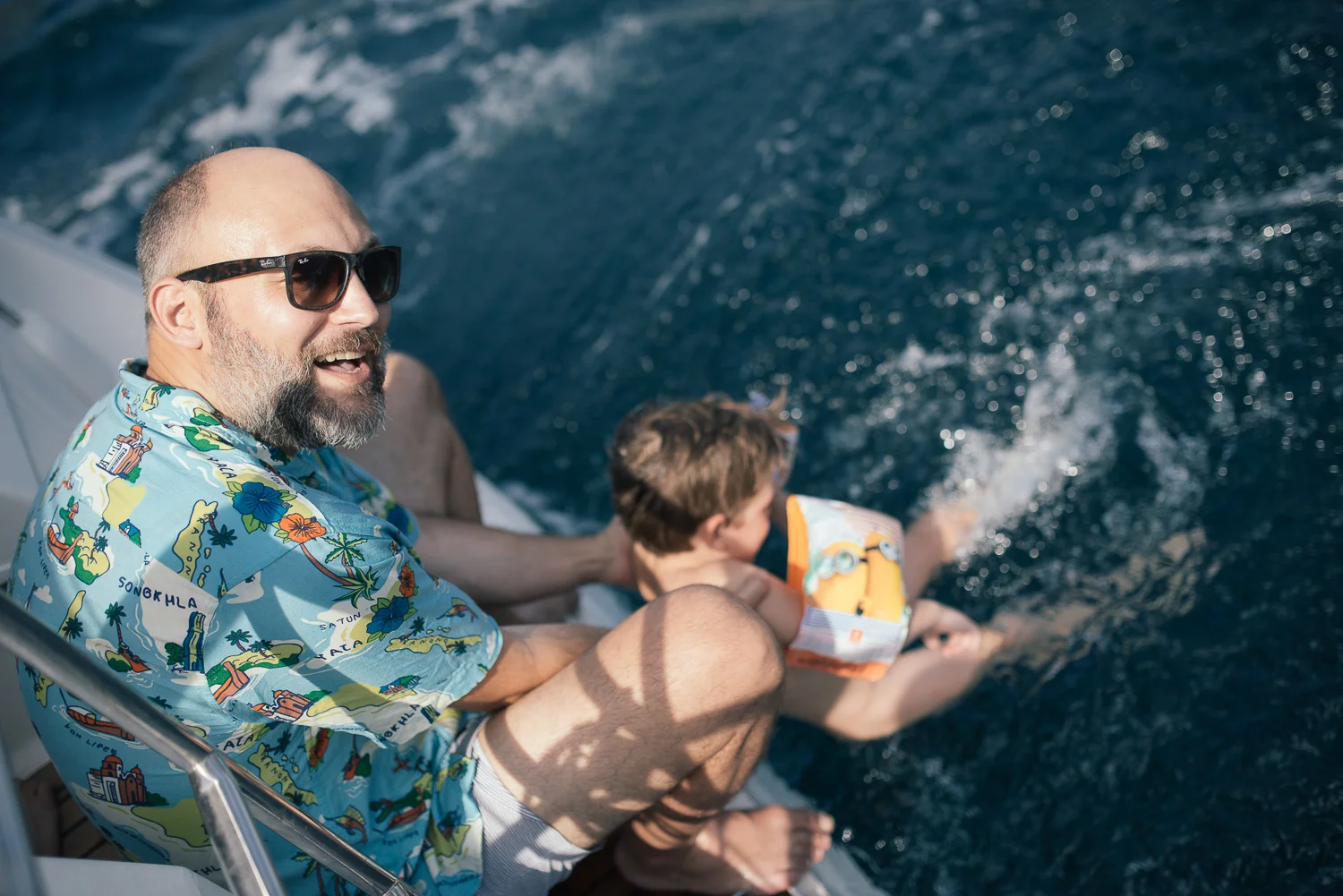 A man with a beard and sunglasses wearing a colorful Hawaiian shirt, smiling while sitting on a boat with a boy in a life jacket, both leaning over the water, enjoying a day on the ocean.