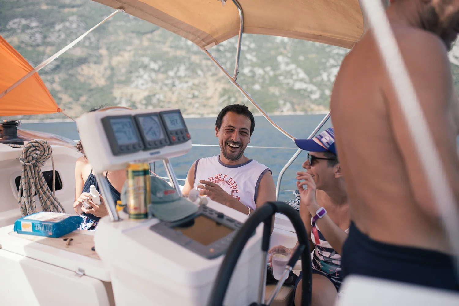 Group of friends sitting on a boat enjoying a sunny day, with a man smiling and others talking, water and hills visible in the background.