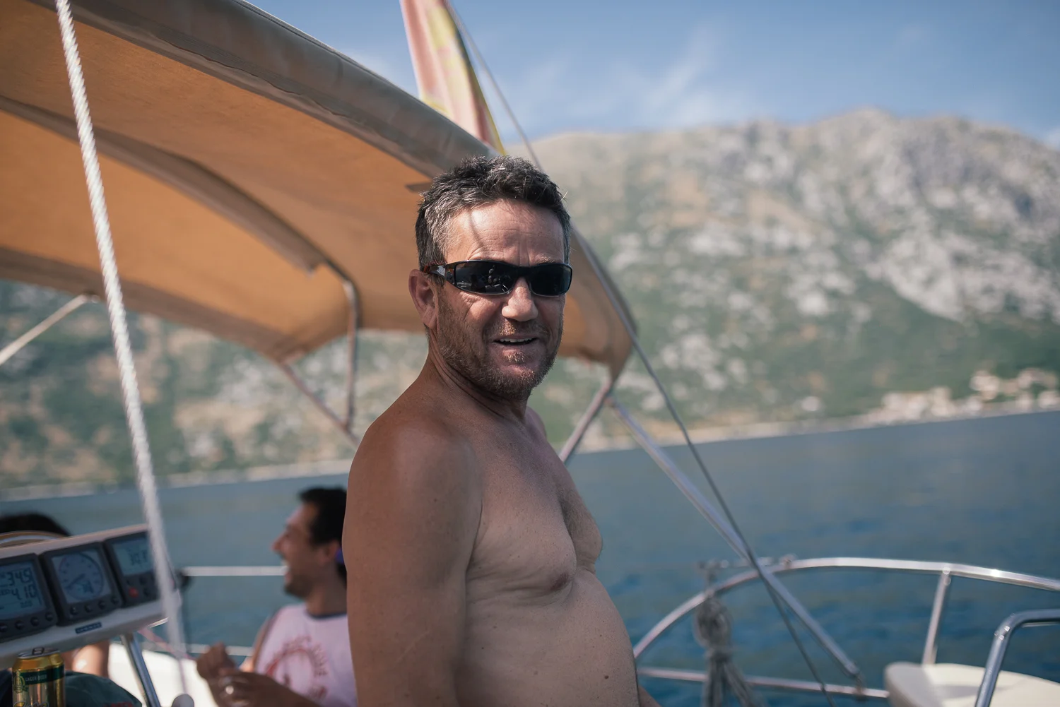 A shirtless man wearing sunglasses smiling on a boat with a man in the background, body of water, and mountains in the distance.