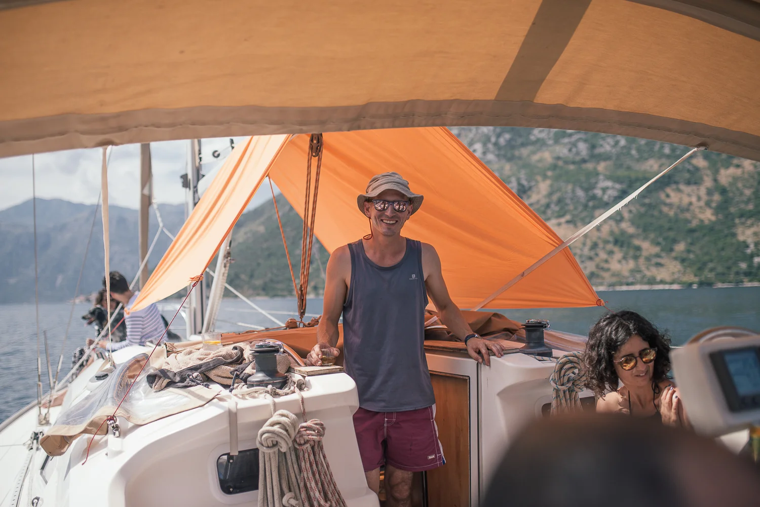 A man with sunglasses, a hat, and a sleeveless shirt standing on a boat, smiling and holding a drink. There is a woman with sunglasses sitting nearby, and another person in the background. The boat is on a body of water with mountains in the distance