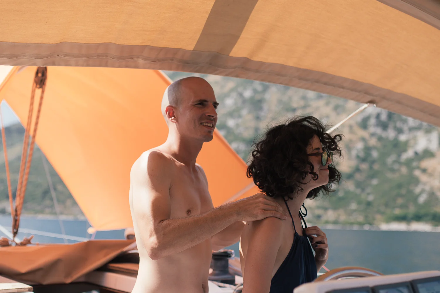 A shirtless man with a shaved head and a woman with curly dark hair and glasses on a boat with beige and orange sail covers, surrounded by water and mountains in the background.