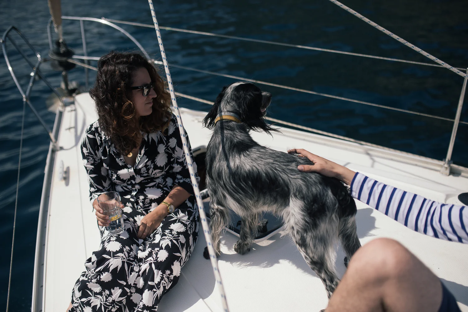 A woman with dark curly hair wearing a black and white floral dress and sunglasses sitting on a boat, holding a glass of water, with a black and white dog next to her, on the deck of a sailboat on the water.