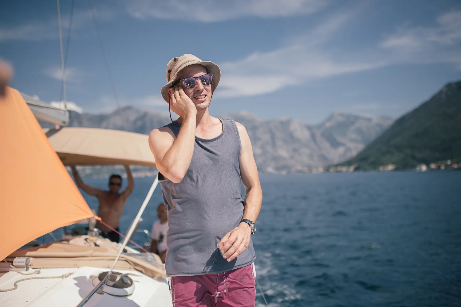A man talking on a cellphone while on a boat, with mountains and water in the background. He is wearing sunglasses, a sun hat, a sleeveless shirt, and shorts. Other people are visible in the background, enjoying a day on the water.