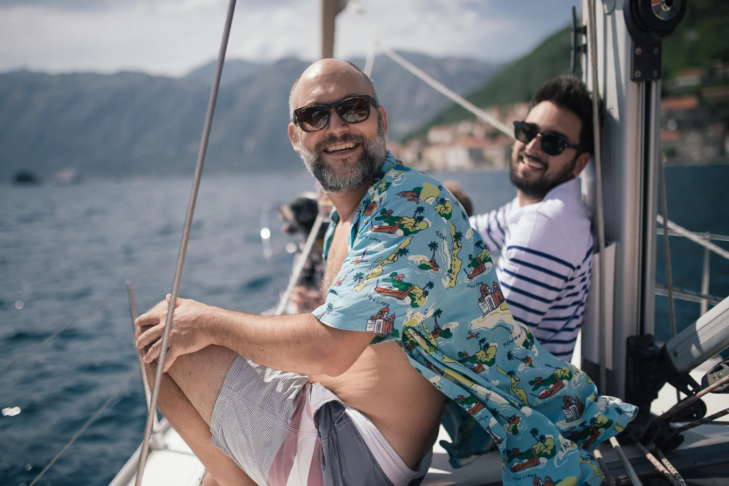 Two men on a boat smiling and enjoying themselves. One is wearing a colorful Hawaiian shirt and sunglasses, while the other is wearing a striped shirt and sunglasses. The background shows a body of water and a distant shoreline with hills.