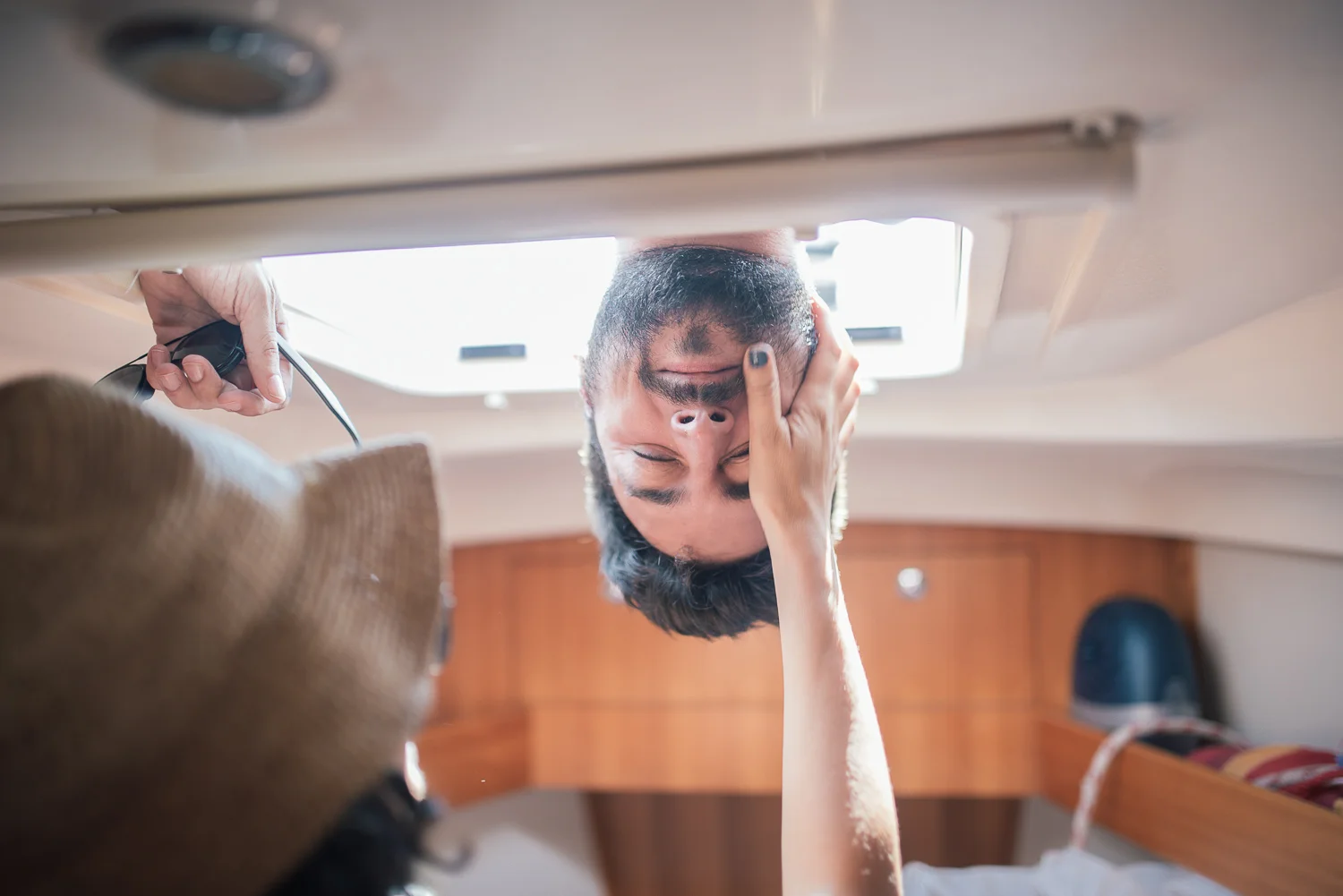 A person with a hat, possibly a hairstylist, holding a device and working on a man's hair or scalp while he rests on his bed with a hand on his forehead, inside a boat cabin.