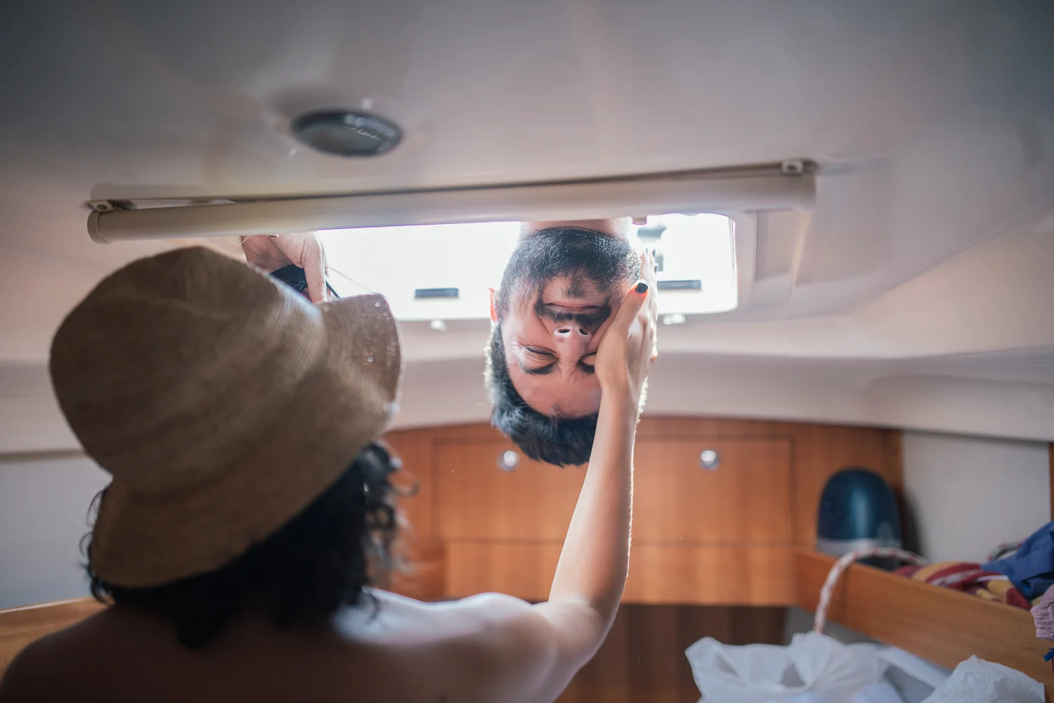 A woman with curly hair and a straw hat touching the face of a man with a beard, who is lying down and opening a porthole in a boat.