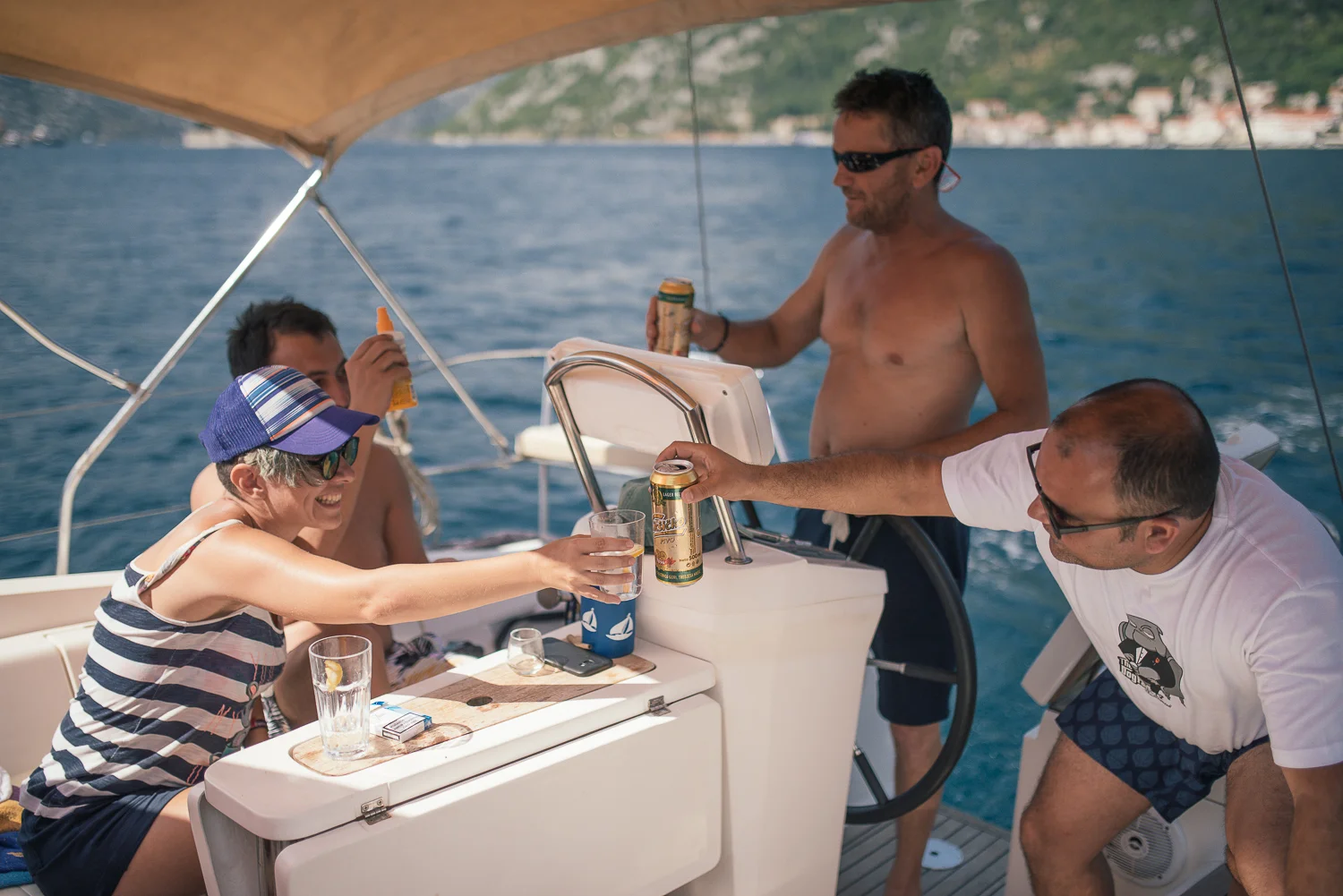 Four friends on a boat enjoying drinks and snacks, with a scenic water and hillside background.