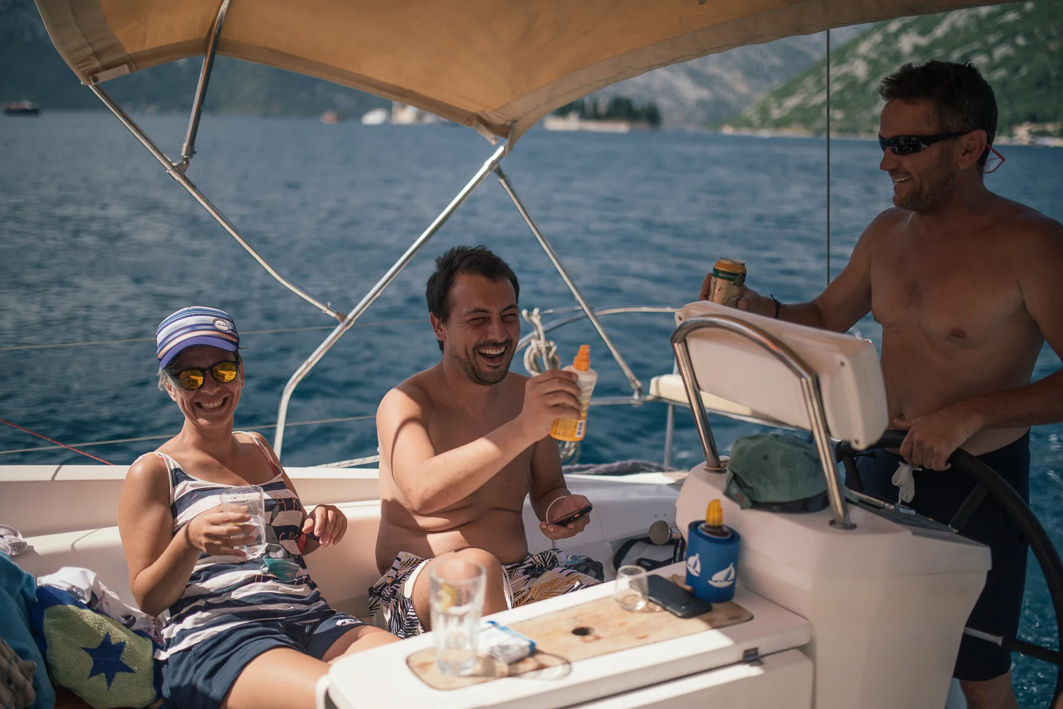Four adults enjoying drinks on a boat in a sunny, scenic lake with cliffs in the background.