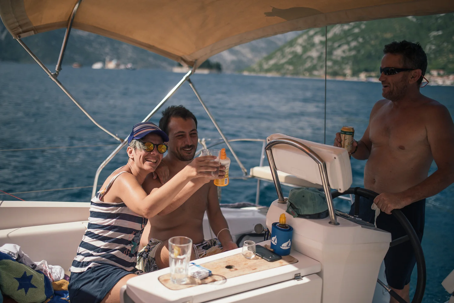 Three people enjoying drinks on a boat with blue water and mountains in the background.