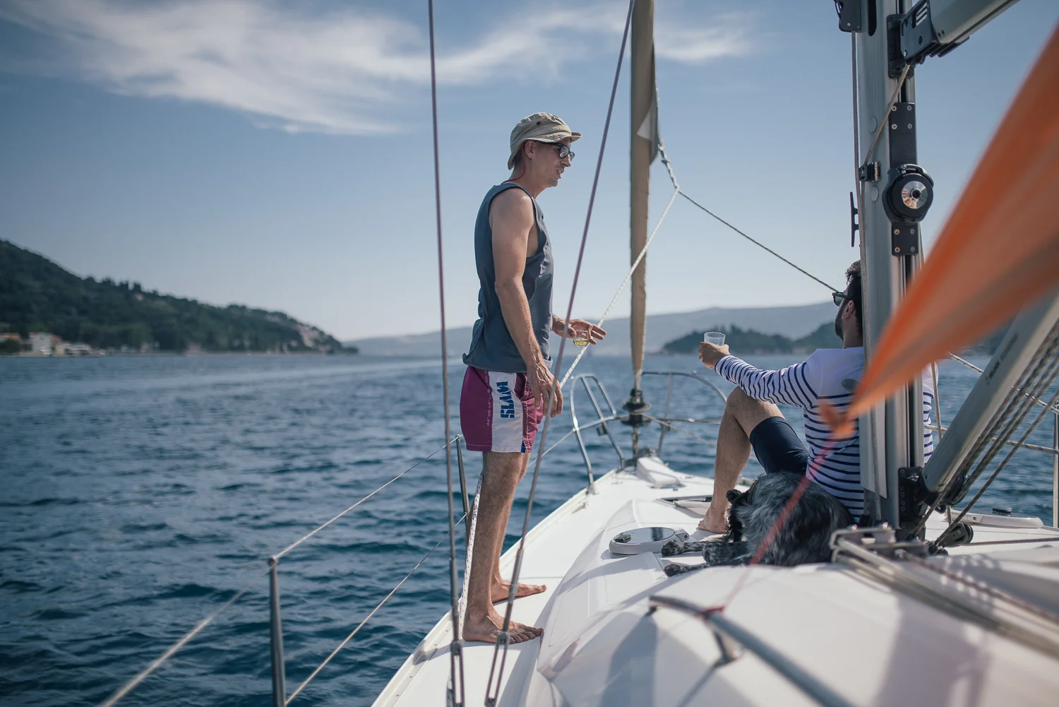 Two men on a sailboat enjoying a sunny day on the water, one is standing and the other is sitting, both with drinks, with a dog lying on the deck.